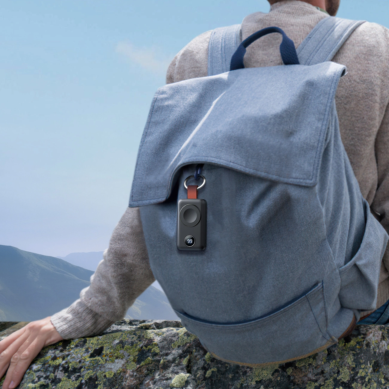 Eine Person sitzt mit Rucksack auf einem Felsen mit Blick auf Berge. Ein schwarzes Gerät hängt am Rucksack und zeigt die Zahl 99.