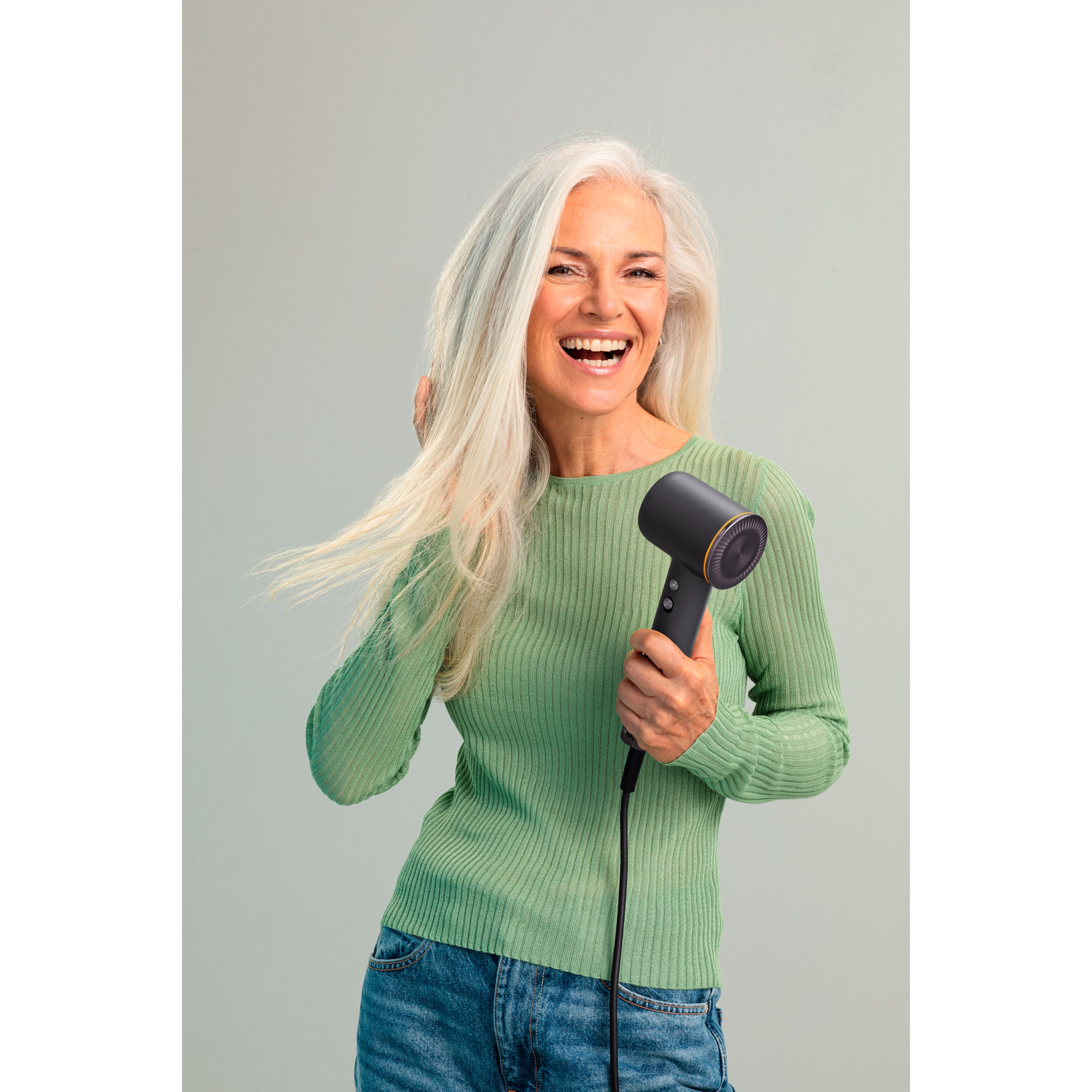 Mujer sonriente con pelo gris sostiene un secador de pelo. Viste suéter verde y jeans. Fondo gris claro.