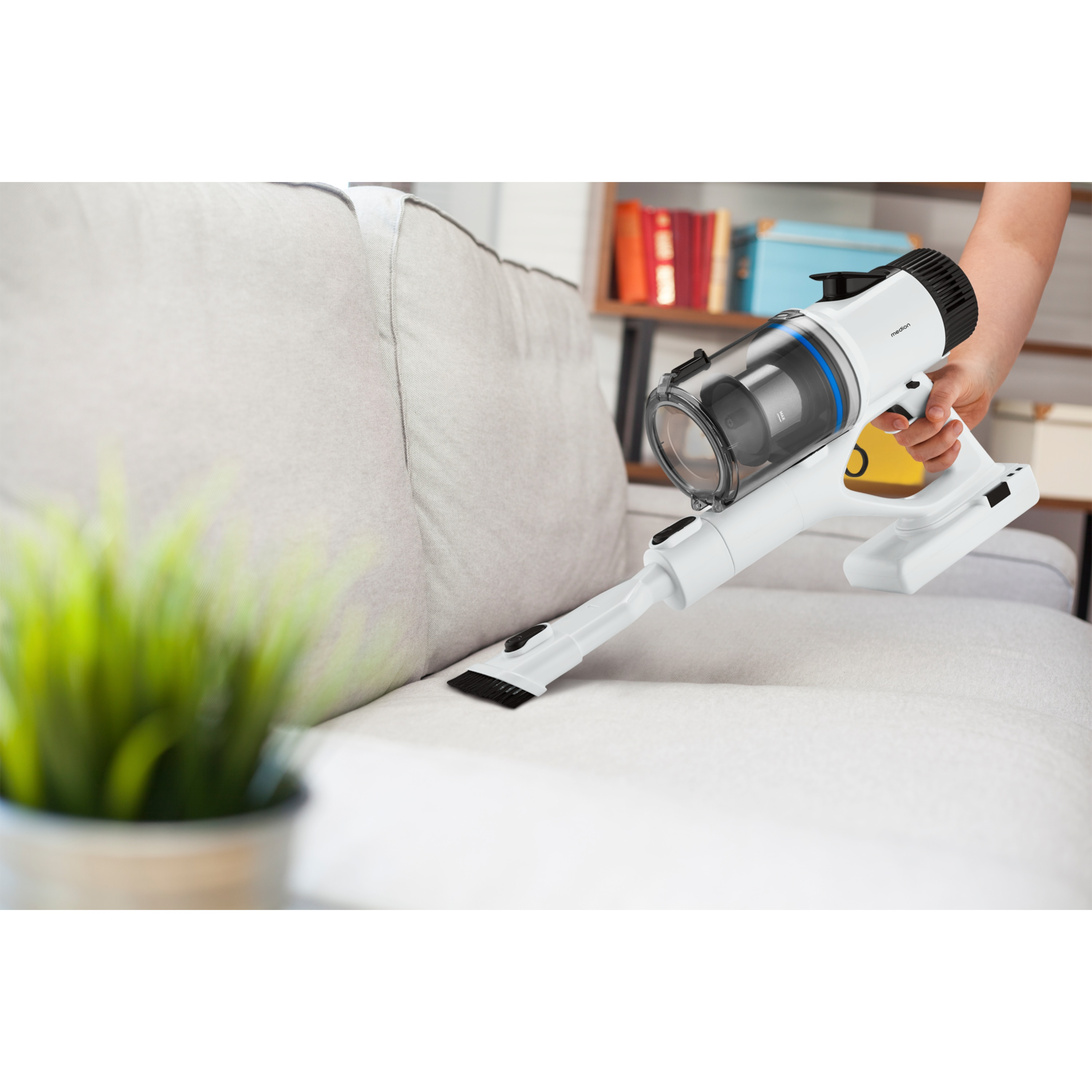 Un aspirateur sans fil blanc et noir utilisé sur un canapé beige. Une plante est au premier plan.