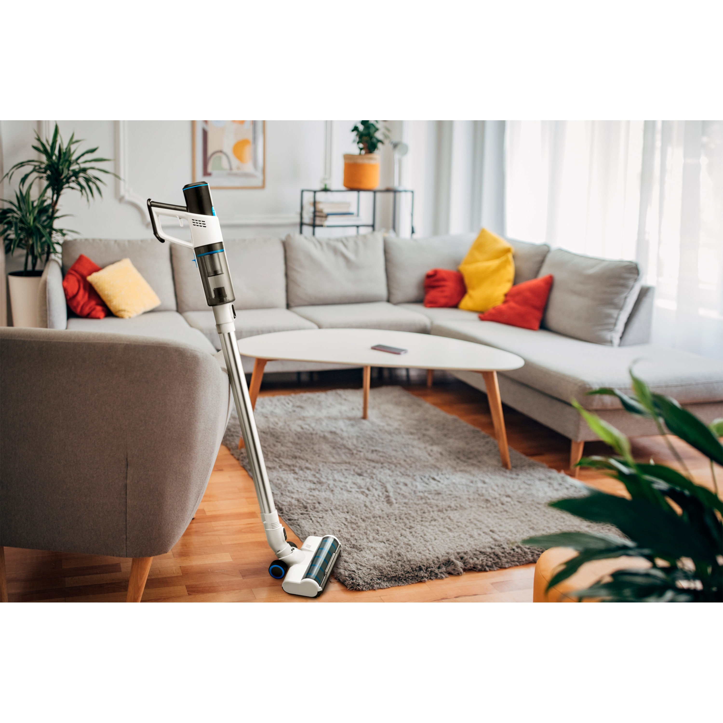 Una aspiradora inalámbrica junto a un sillón gris. Un sofá gris con cojines coloridos en el fondo.