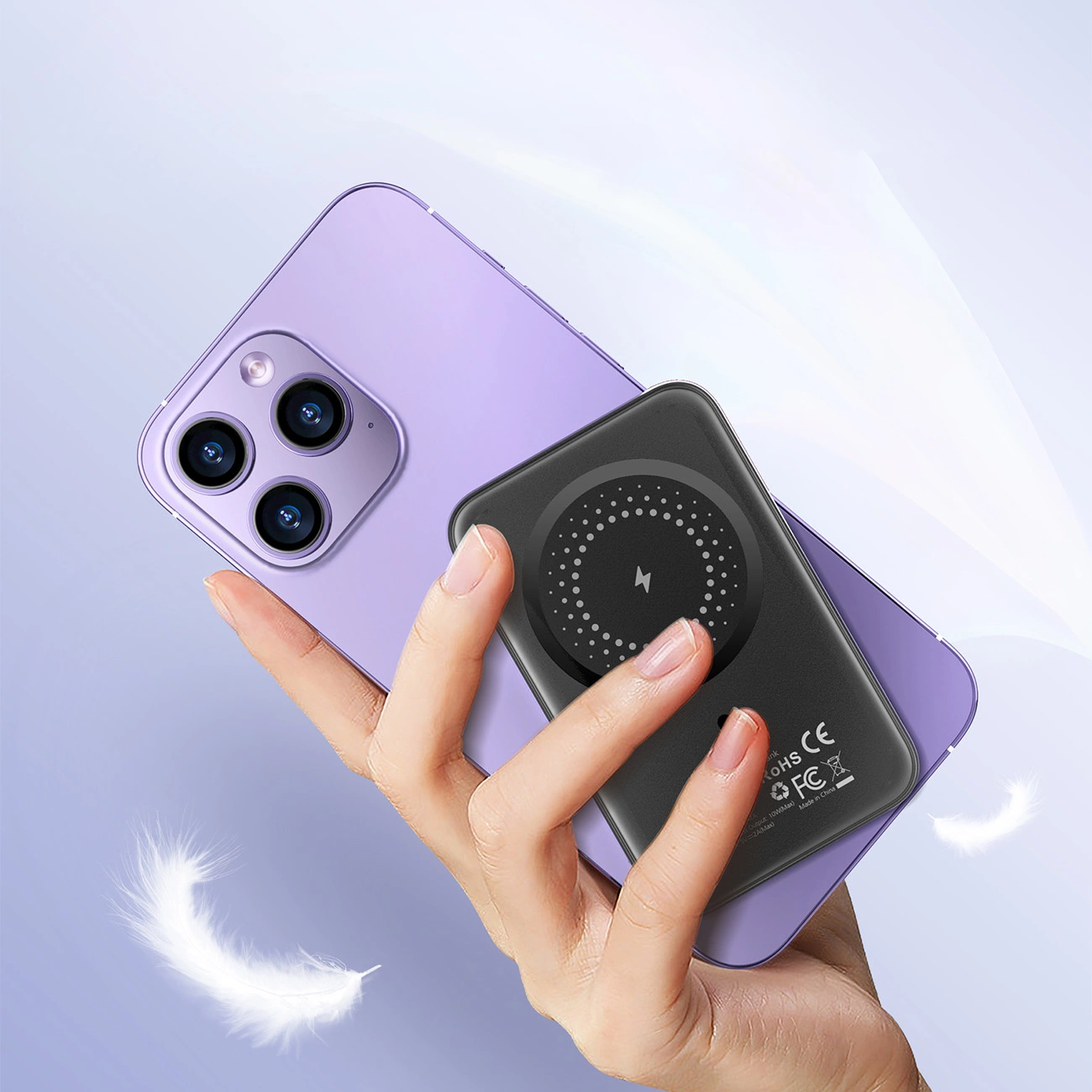 Eine Hand hält ein schwarzes kabelloses Ladegerät auf einem lila Telefon. Federn schweben herum. Fokus auf die Oberfläche des Ladegeräts.