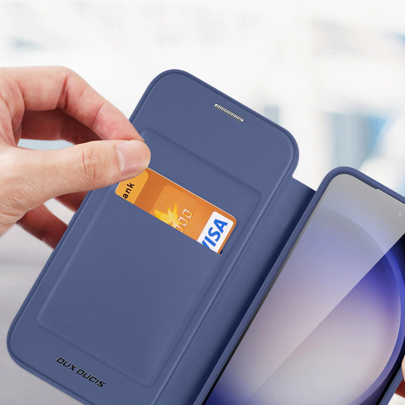 Mano che tiene una custodia blu per telefono con carta di credito nella fessura. Un telefono è visibile a destra. Dux Ducis in basso.