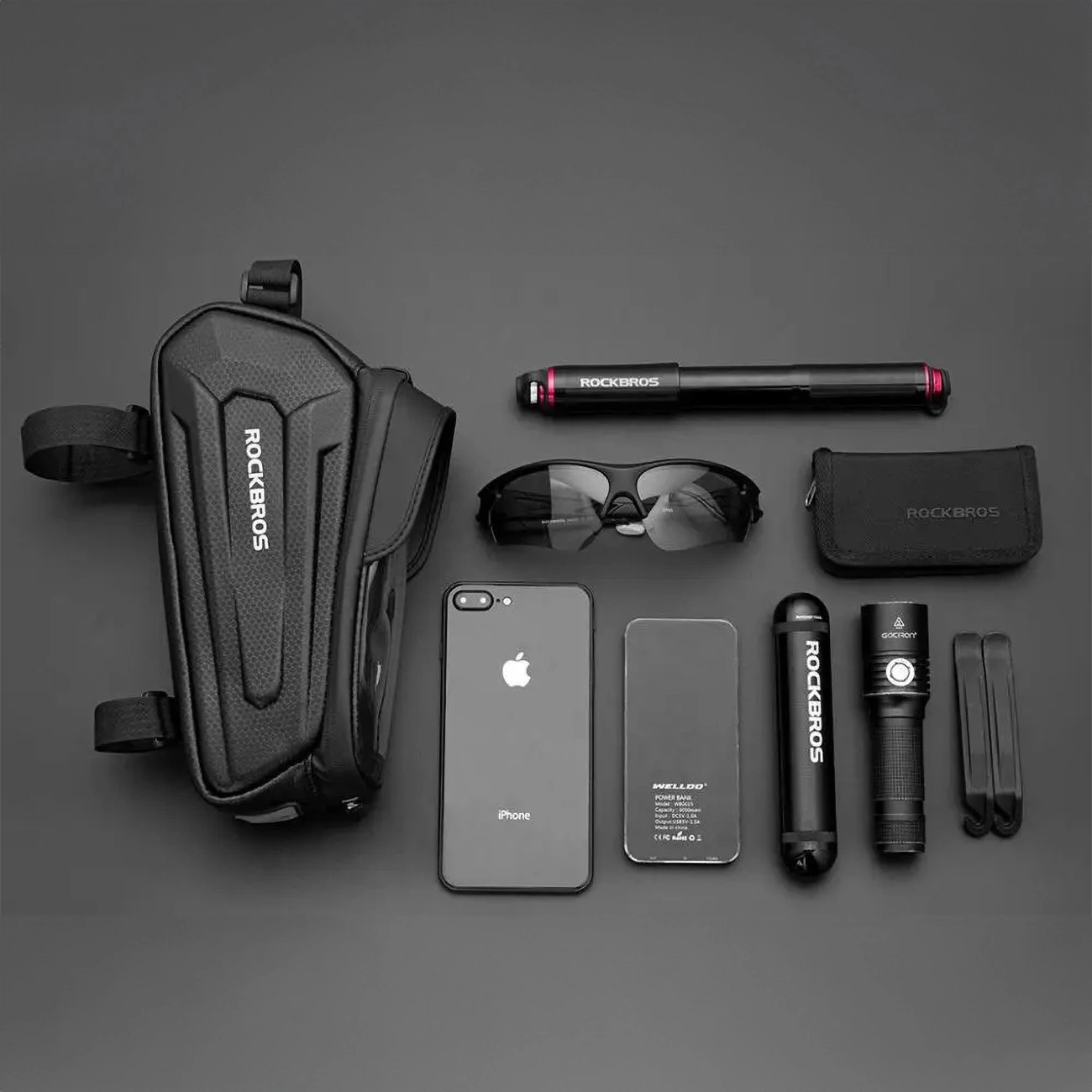 Vista dall'alto di accessori da ciclismo: borsa, pompa, occhiali da sole, custodia, telefono, power bank, torcia e attrezzi, il tutto su una superficie grigia.