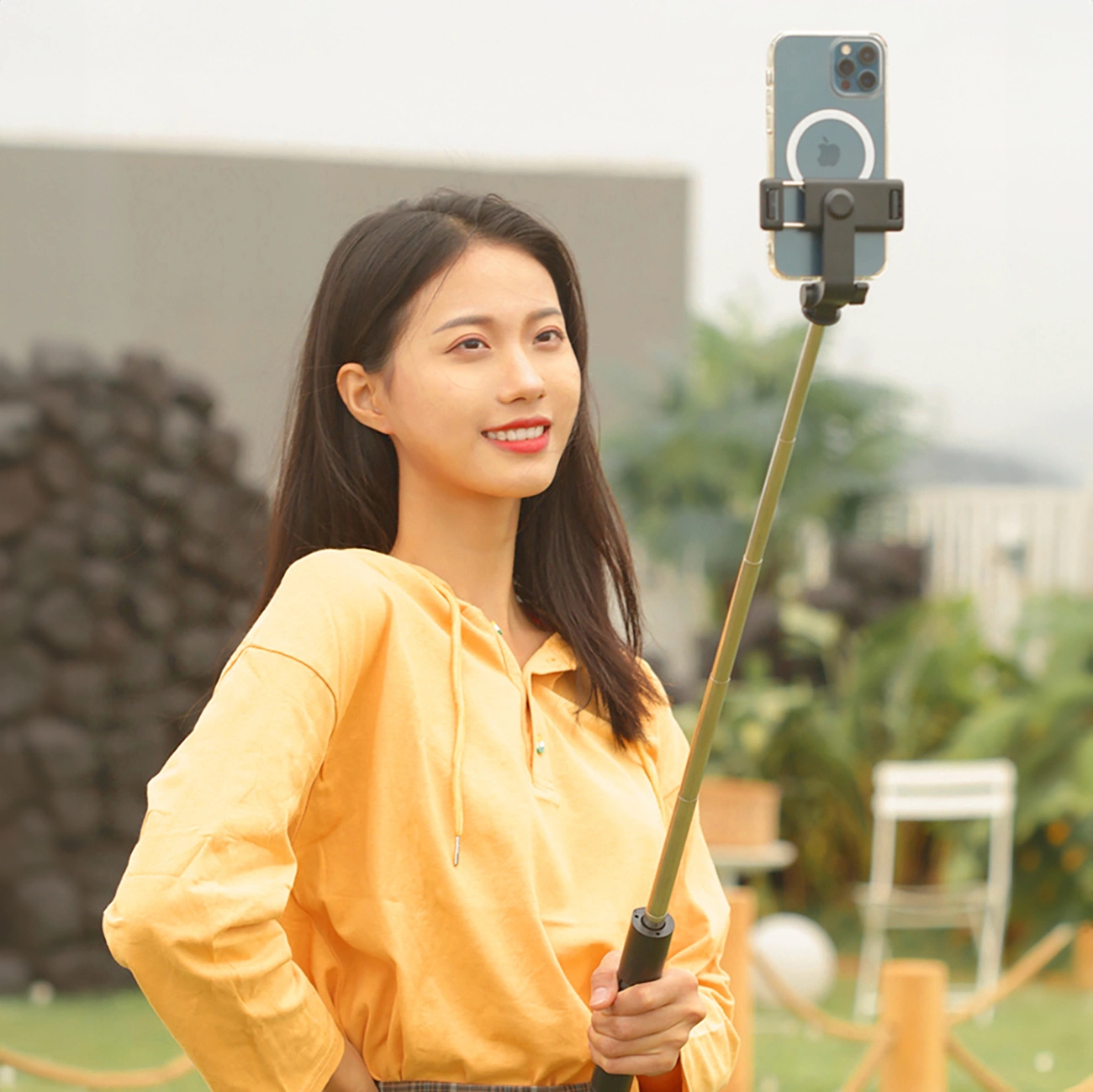 Eine Frau hält einen Selfie-Stick mit einem Telefon. Sie trägt ein gelbes Hemd und lächelt im Freien mit Grün im Hintergrund.