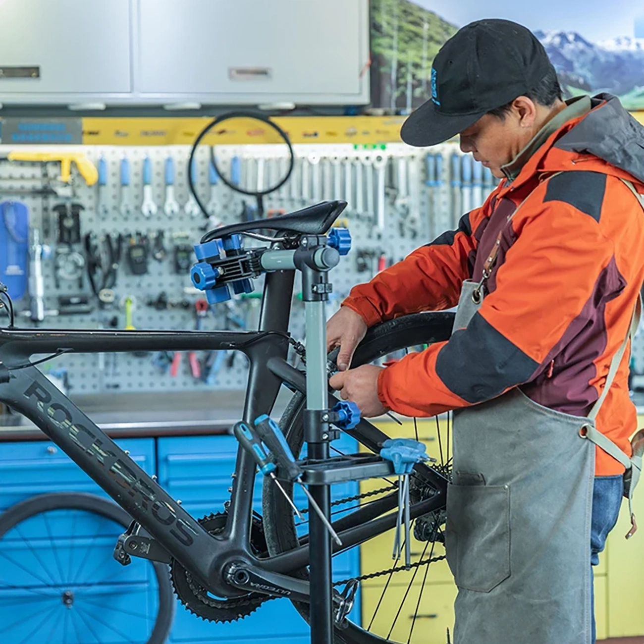 Ein Mann in einer orangefarbenen Jacke repariert ein Fahrrad. Das Fahrrad steht in einer Werkstatt mit Werkzeugen an der Wand.