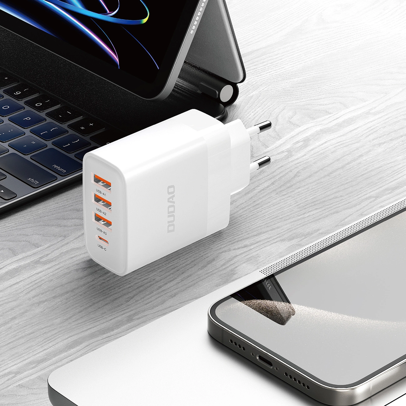 Biała ładowarka DUDO z wieloma portami USB znajduje się na drewnianej powierzchni, w pobliżu laptopa i telefonu.