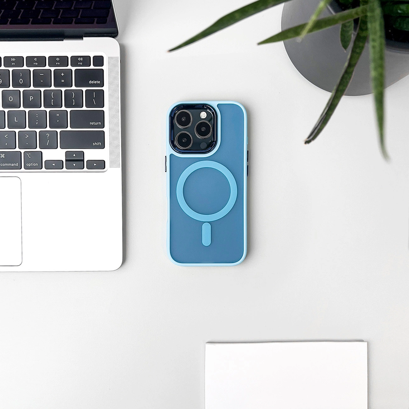 Una custodia blu per telefono con un design circolare e un iPhone si trova su una superficie bianca vicino a un laptop e una pianta.