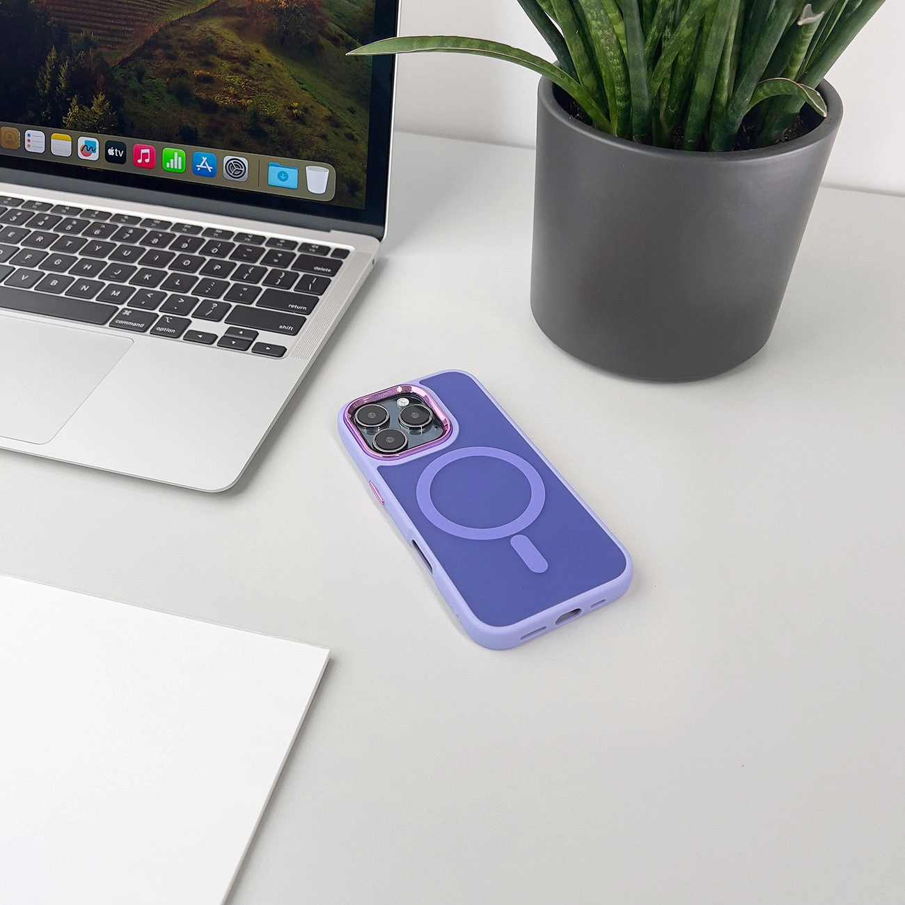 Un laptop, una custodia viola con un cerchio, una pianta e un foglio bianco su un tavolo bianco. Lo sfondo mostra un paesaggio.