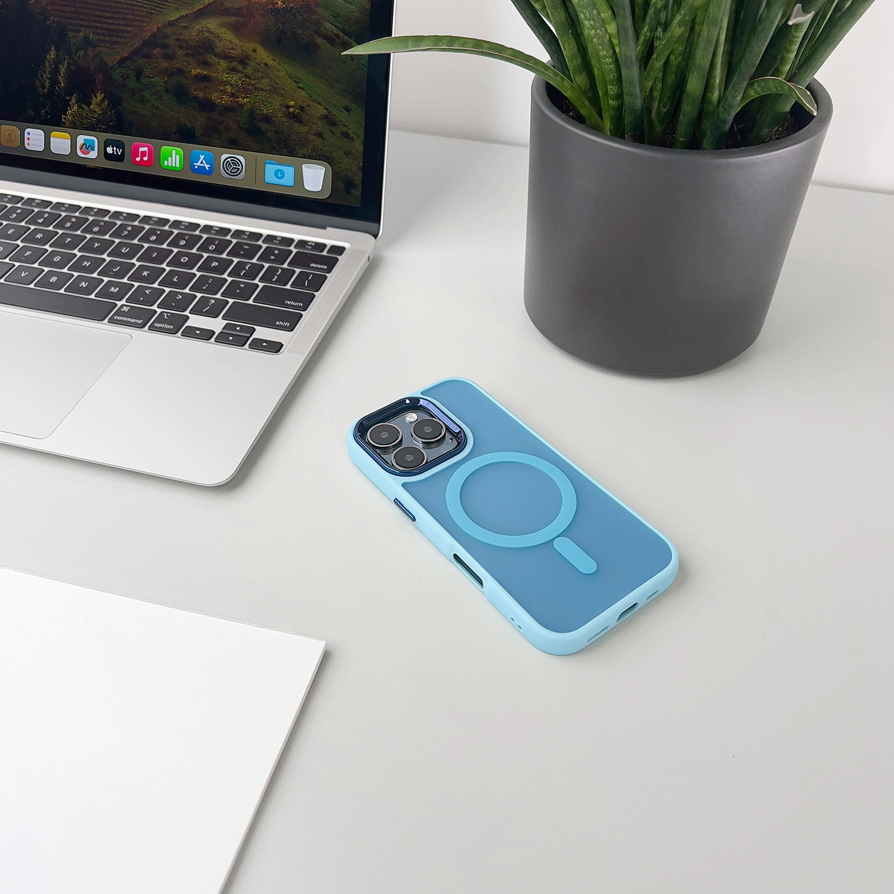 Un laptop, una custodia blu per telefono, una pianta in un vaso scuro e un foglio bianco su una scrivania bianca sono visibili.