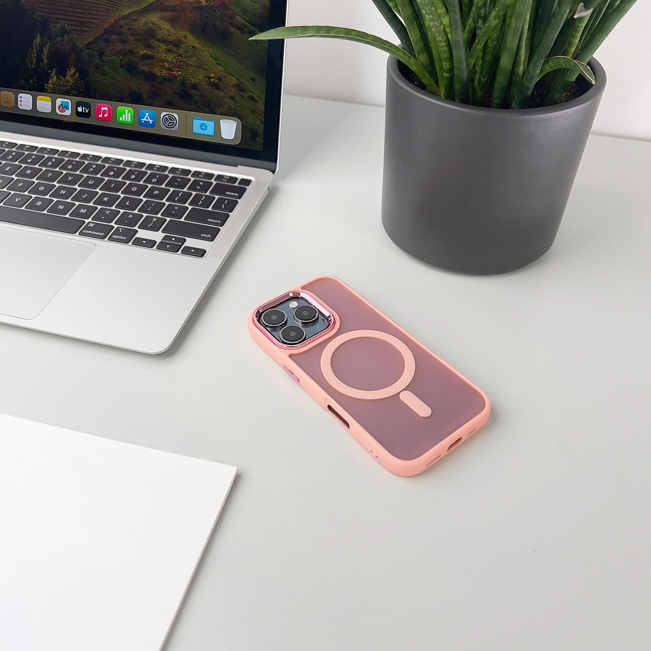 Un laptop, una custodia per telefono e una pianta in vaso sono su una scrivania bianca. La custodia ha una cornice rosa e un centro rosa chiaro.