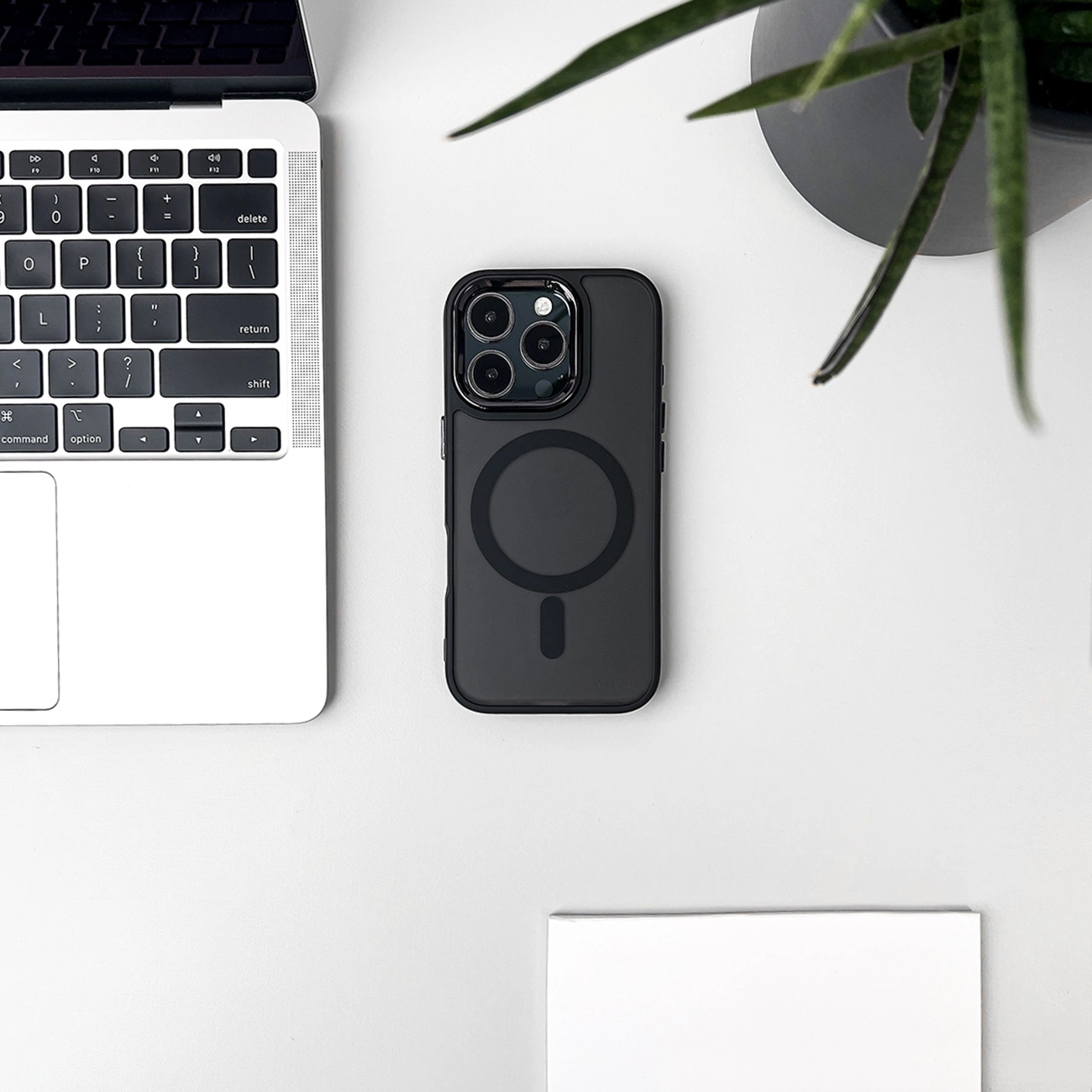 Una custodia nera per telefono con un cerchio magnetico e una tastiera per laptop sono su una superficie bianca. Una pianta sullo sfondo.