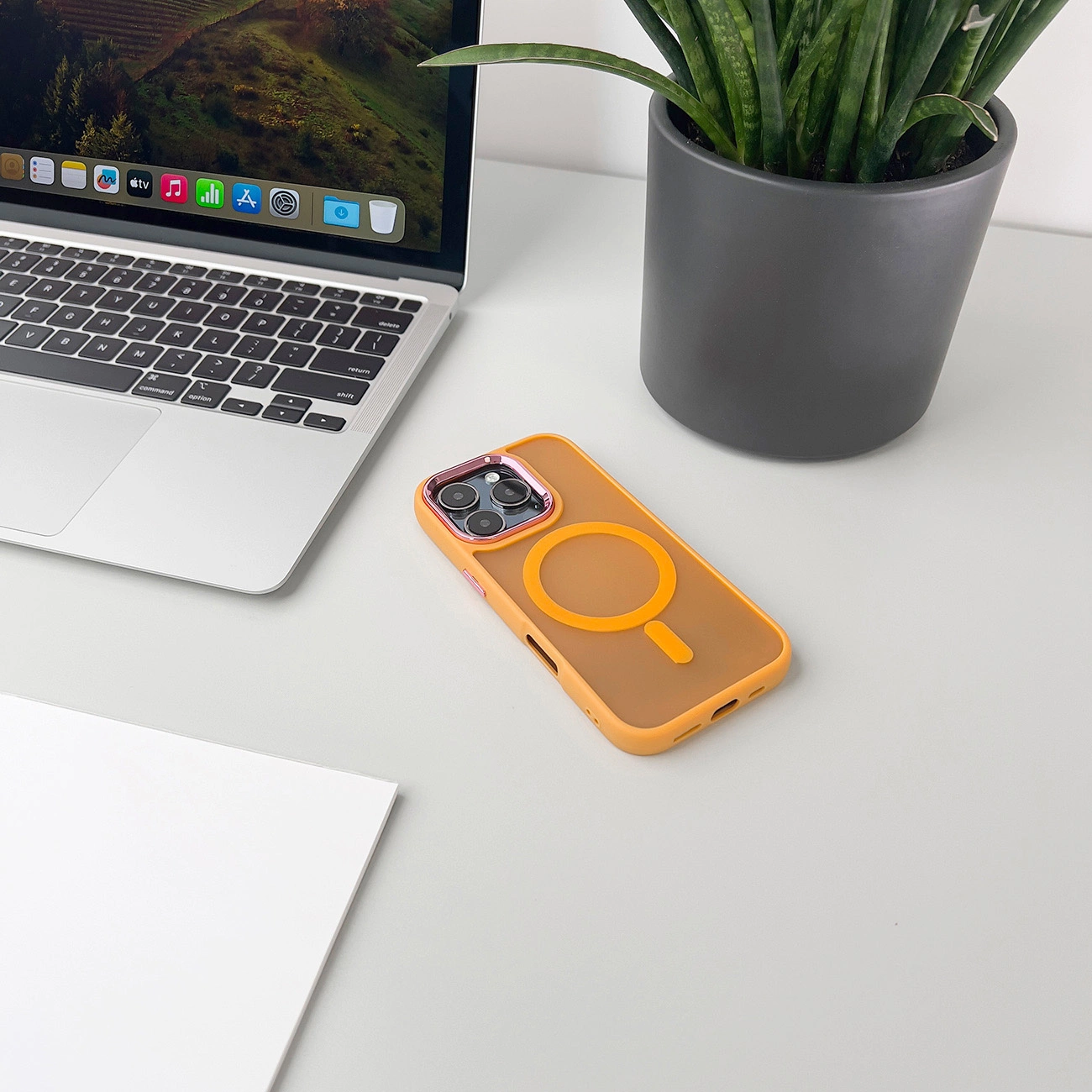 Un laptop, una custodia per telefono e una pianta su una scrivania bianca. La custodia del telefono è arancione con un design circolare.
