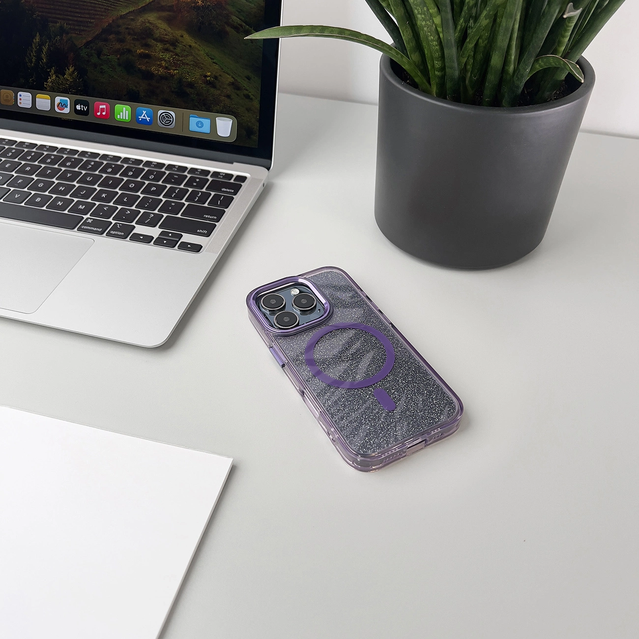 Un laptop, una custodia per telefono e una pianta su una scrivania bianca. La custodia del telefono è viola e argento. Il laptop mostra un paesaggio.