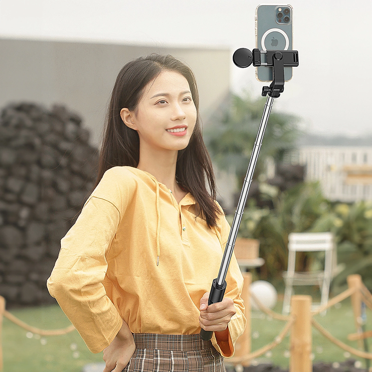 Eine Frau hält einen Selfie-Stick mit einem Telefon. Sie trägt ein gelbes Hemd und einen karierten Rock und lächelt im Freien.