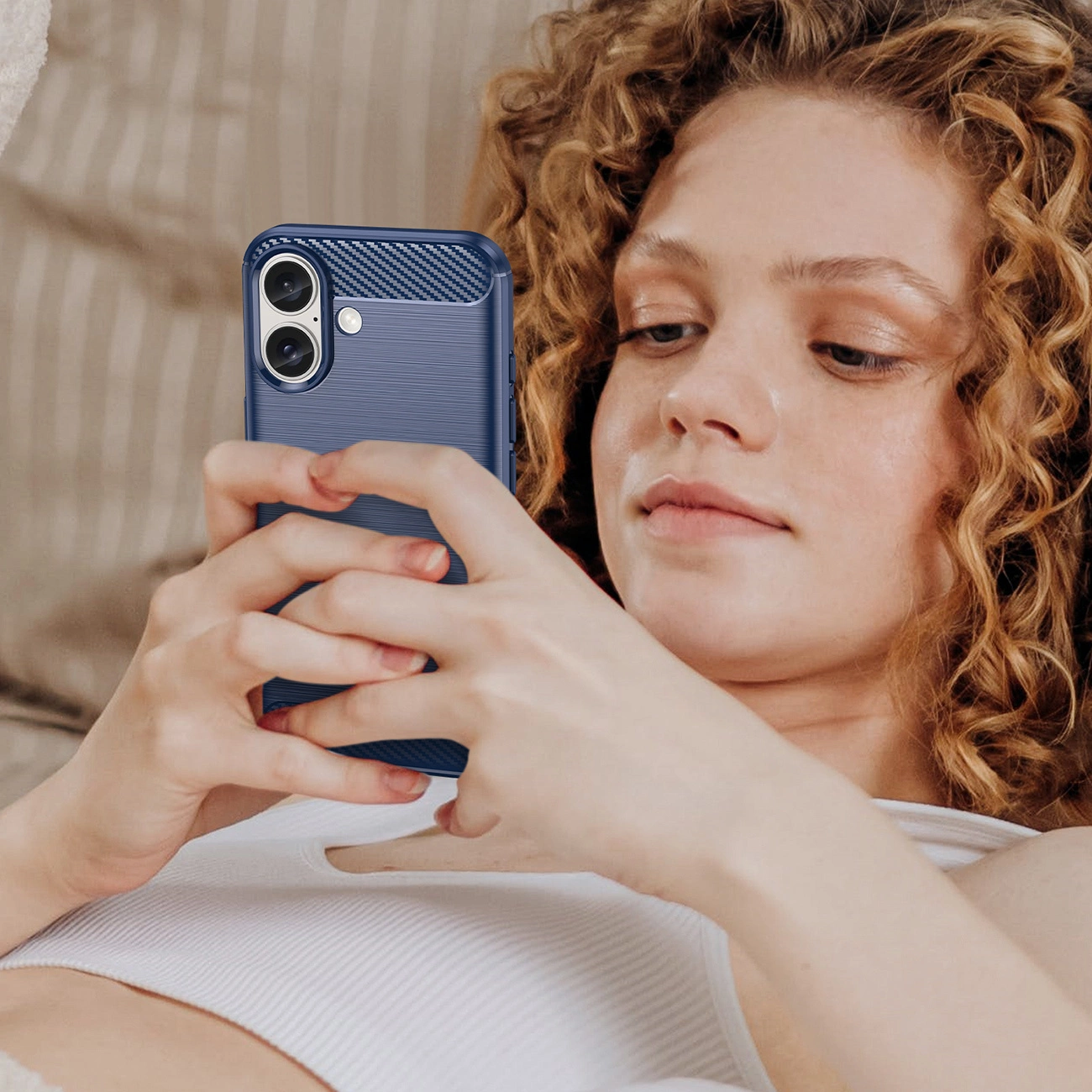 Una donna con i capelli ricci tiene in mano un telefono blu. Indossa un top bianco. Lo sfondo è beige sfocato.