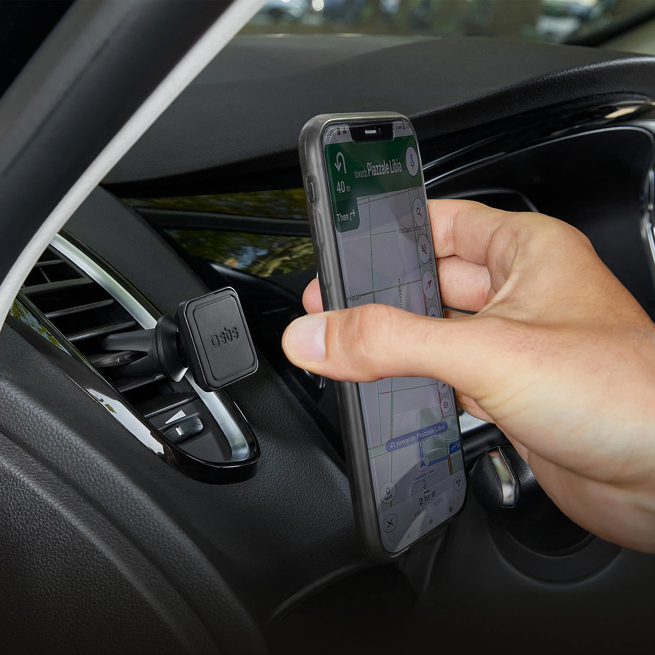 Una mano tiene in mano un telefono che mostra una mappa in un'auto. Il telefono è montato su una bocchetta dell'aria del cruscotto. La navigazione mostra Piazzale Libia.