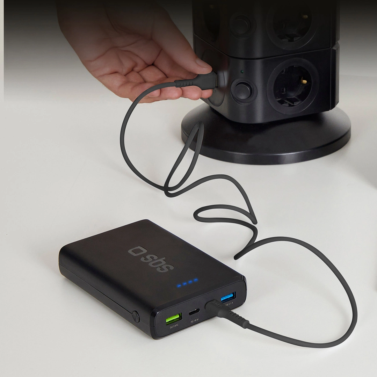 Een hand steekt een zwarte kabel in een zwarte stroomtoren. Een zwarte powerbank met een aangesloten kabel ligt op een wit oppervlak.