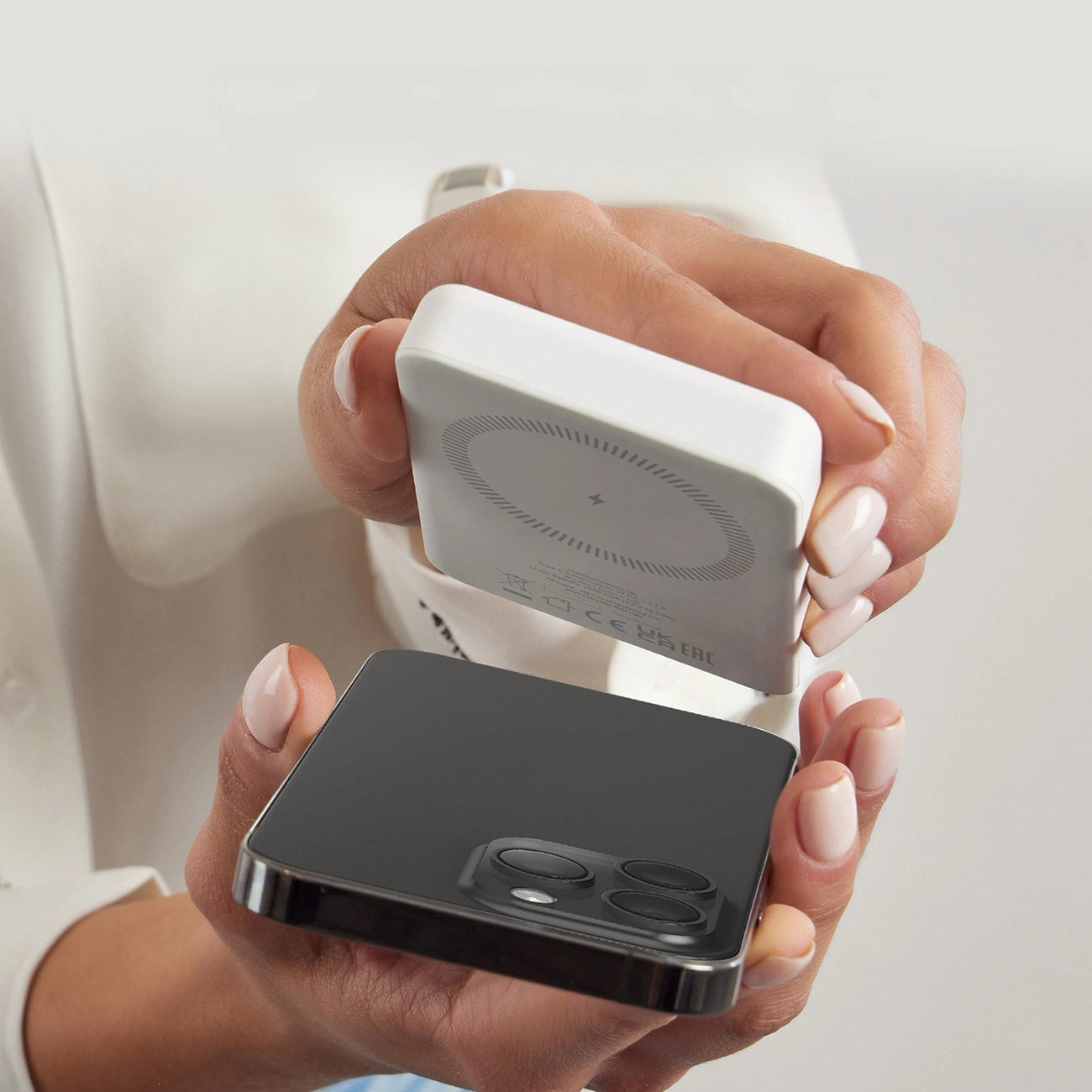 Mani che tengono un caricabatterie wireless bianco sopra uno smartphone nero. Il caricabatterie ha un simbolo di ricarica. Lo sfondo è bianco.