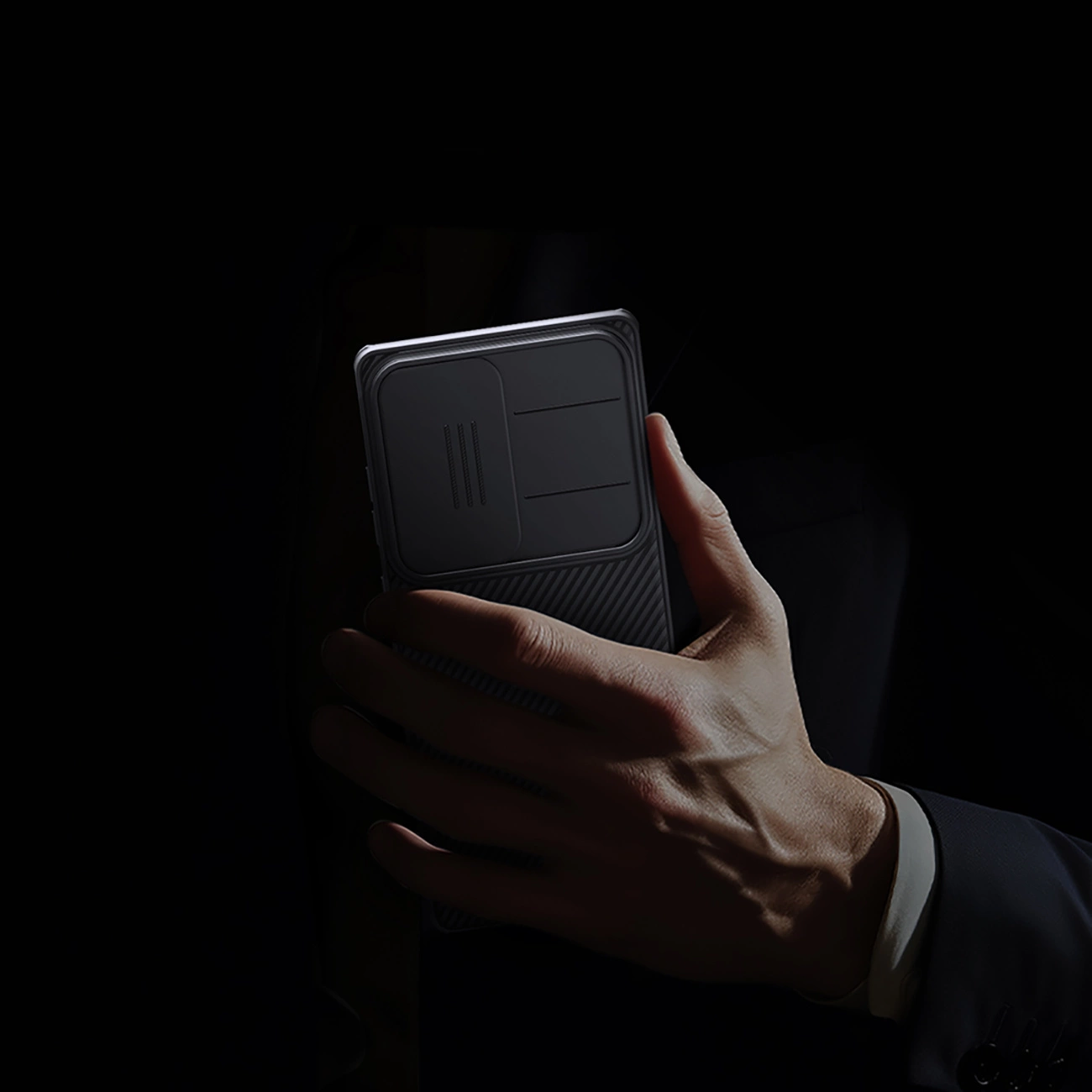 Eine Hand hält ein dunkles Telefon mit strukturierter Rückseite. Die Person trägt einen Anzug. Der Hintergrund ist dunkel, was das Telefon hervorhebt.