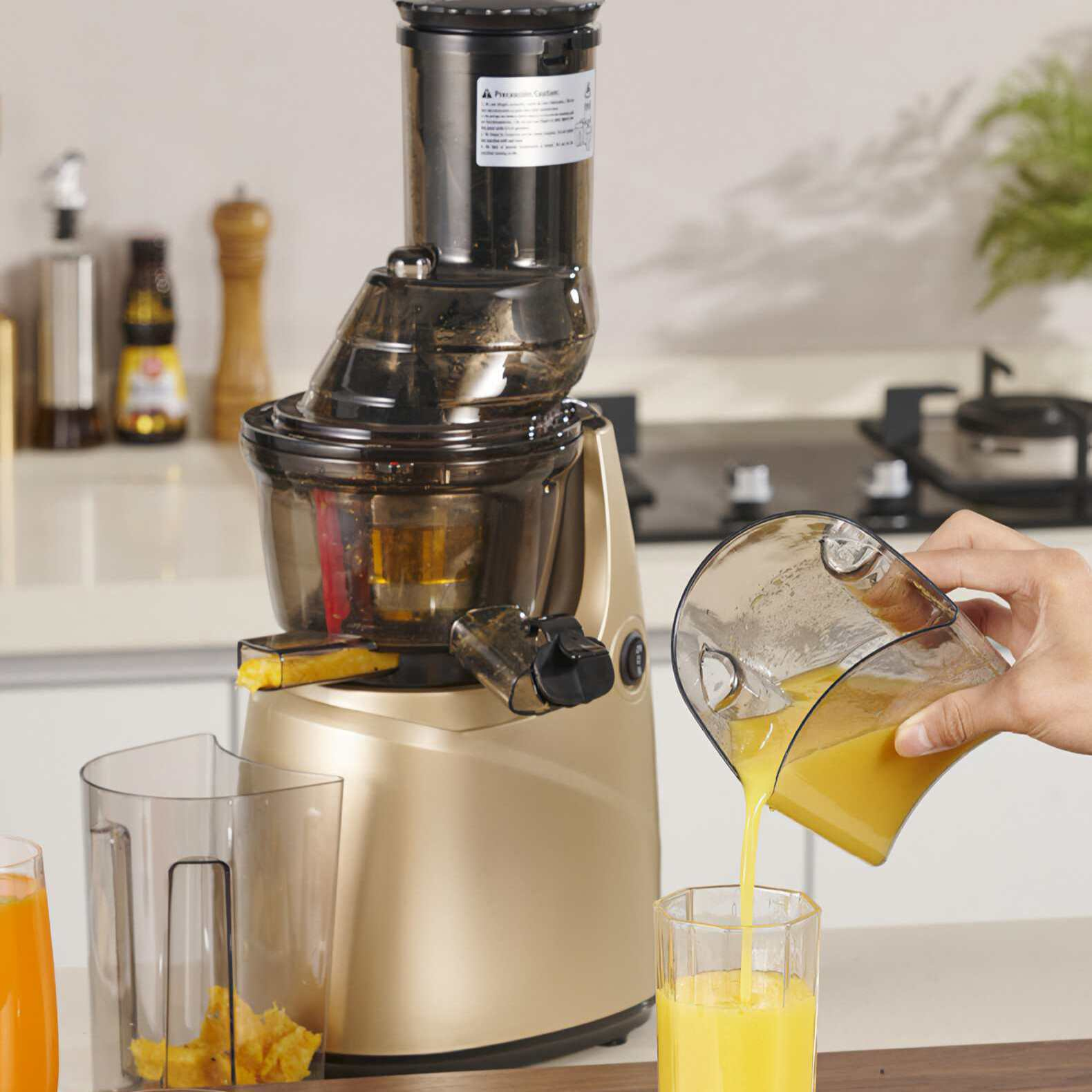 Ein Entsafter macht Orangensaft. Eine Hand gießt Saft aus einem Behälter in ein Glas. Küchenhintergrund.