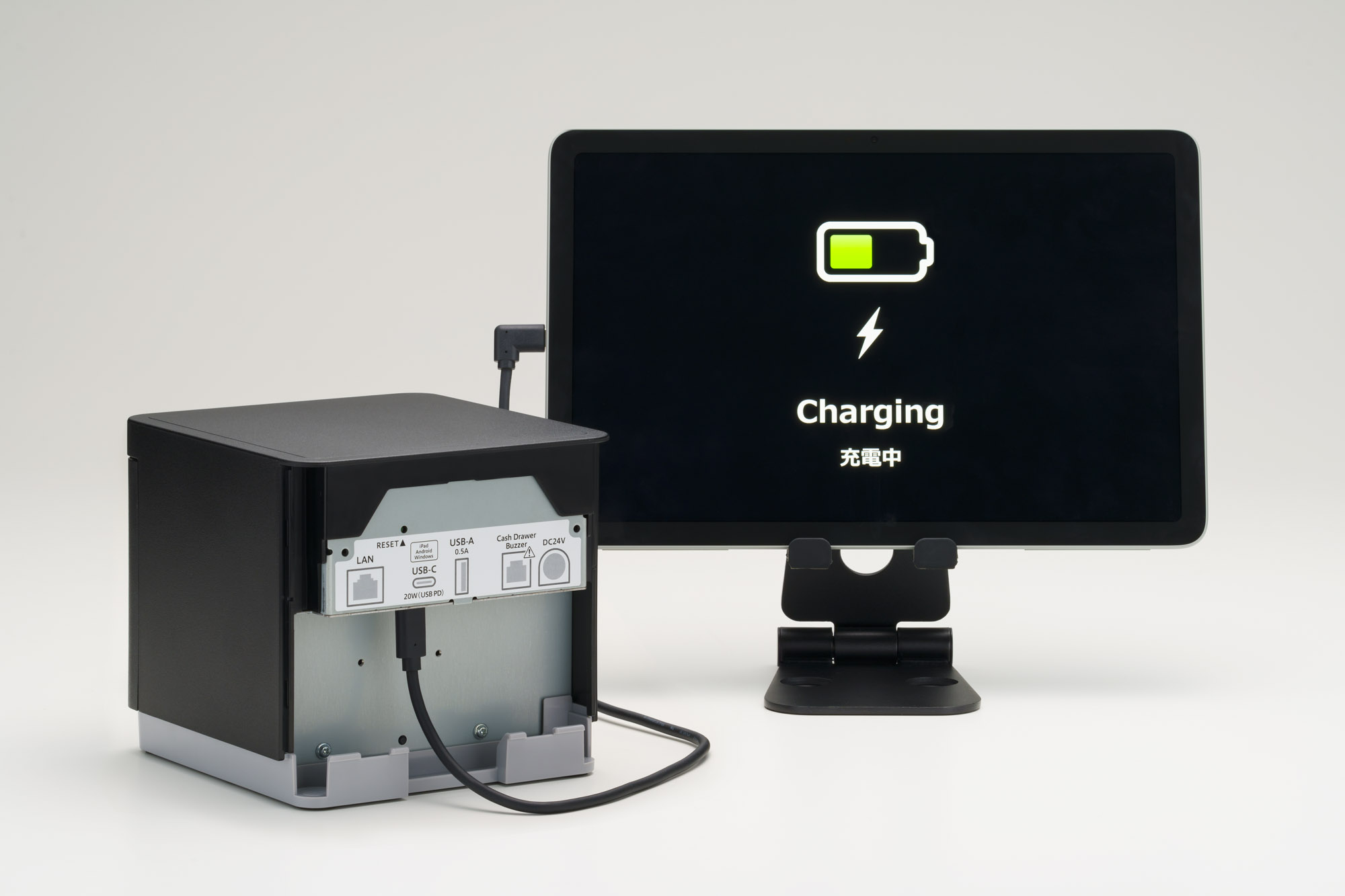 Tablet wyświetla 'Charging'. Po lewej czarne pudełko z portami i kablem ładującym. Stojak podtrzymuje tablet.