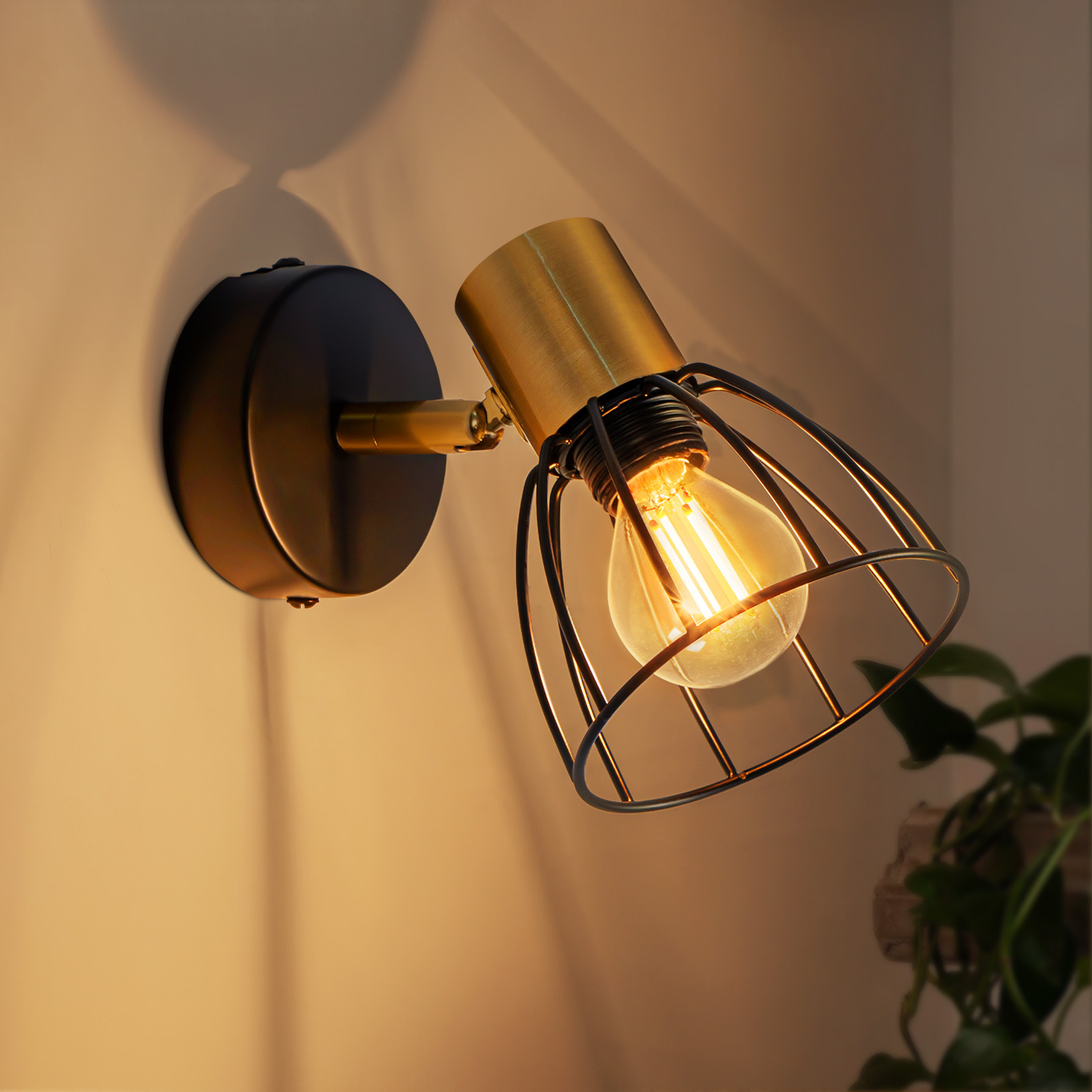 Una lampada nera e dorata a parete con una lampadina a gabbia illumina una parete beige. Una pianta è sullo sfondo.