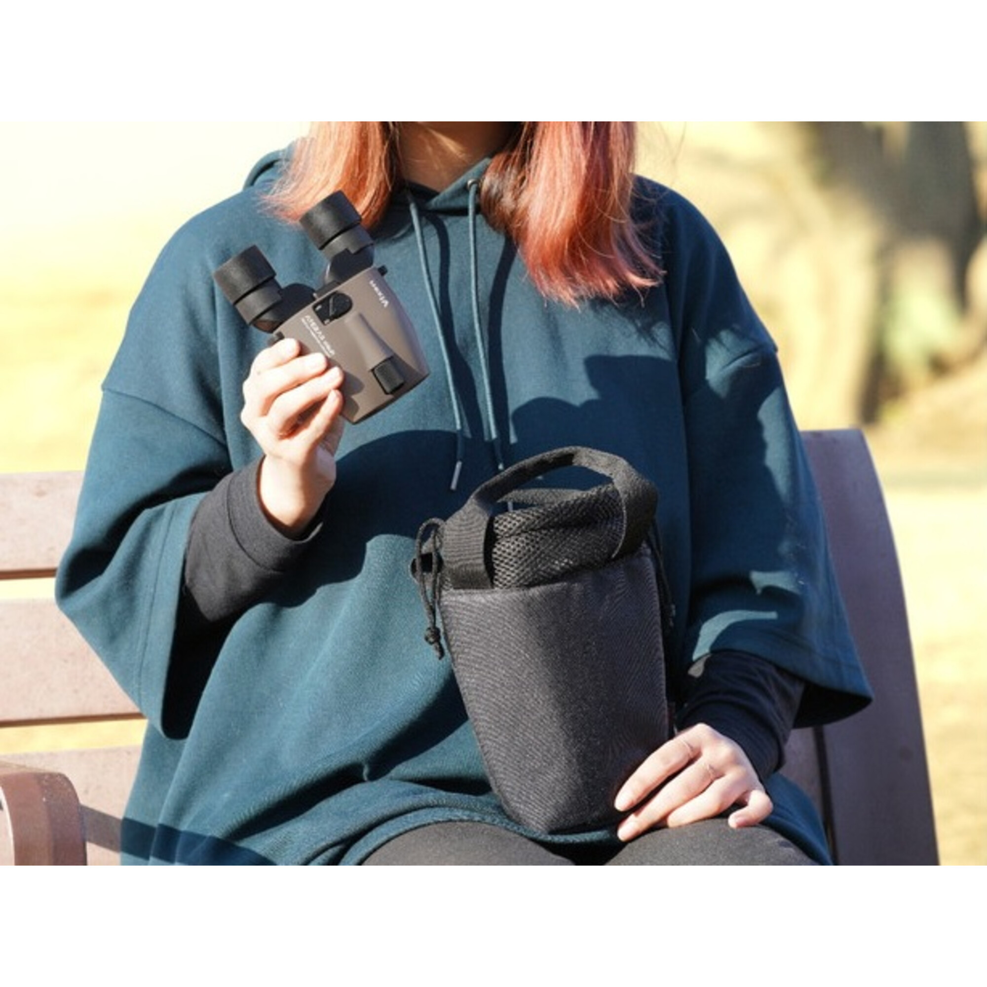 Eine Person in einem teal Hoodie hält ein Fernglas und eine schwarze Tasche und sitzt draußen auf einer Bank.