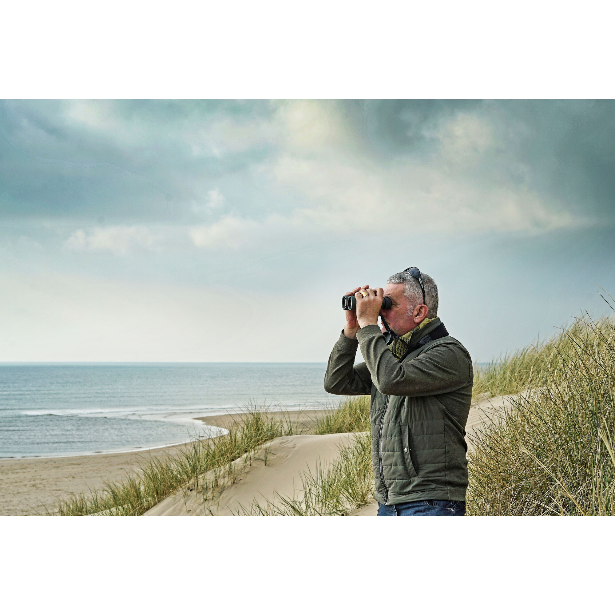 Ein Mann in einer grünen Jacke benutzt ein Fernglas auf einer grasbewachsenen Düne mit Blick auf das Meer.