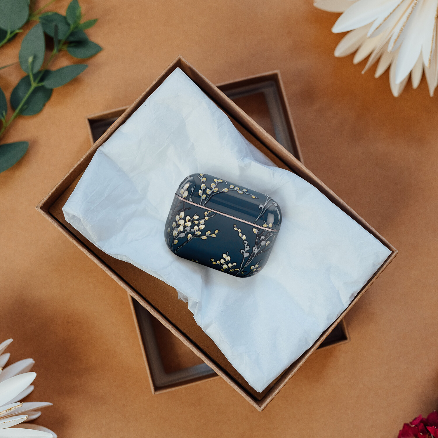 Ein dunkelblaues Etui mit goldenem Blumendesign liegt in einer offenen Geschenkbox. Die Box steht auf einer braunen Oberfläche mit Blumen und Grün.