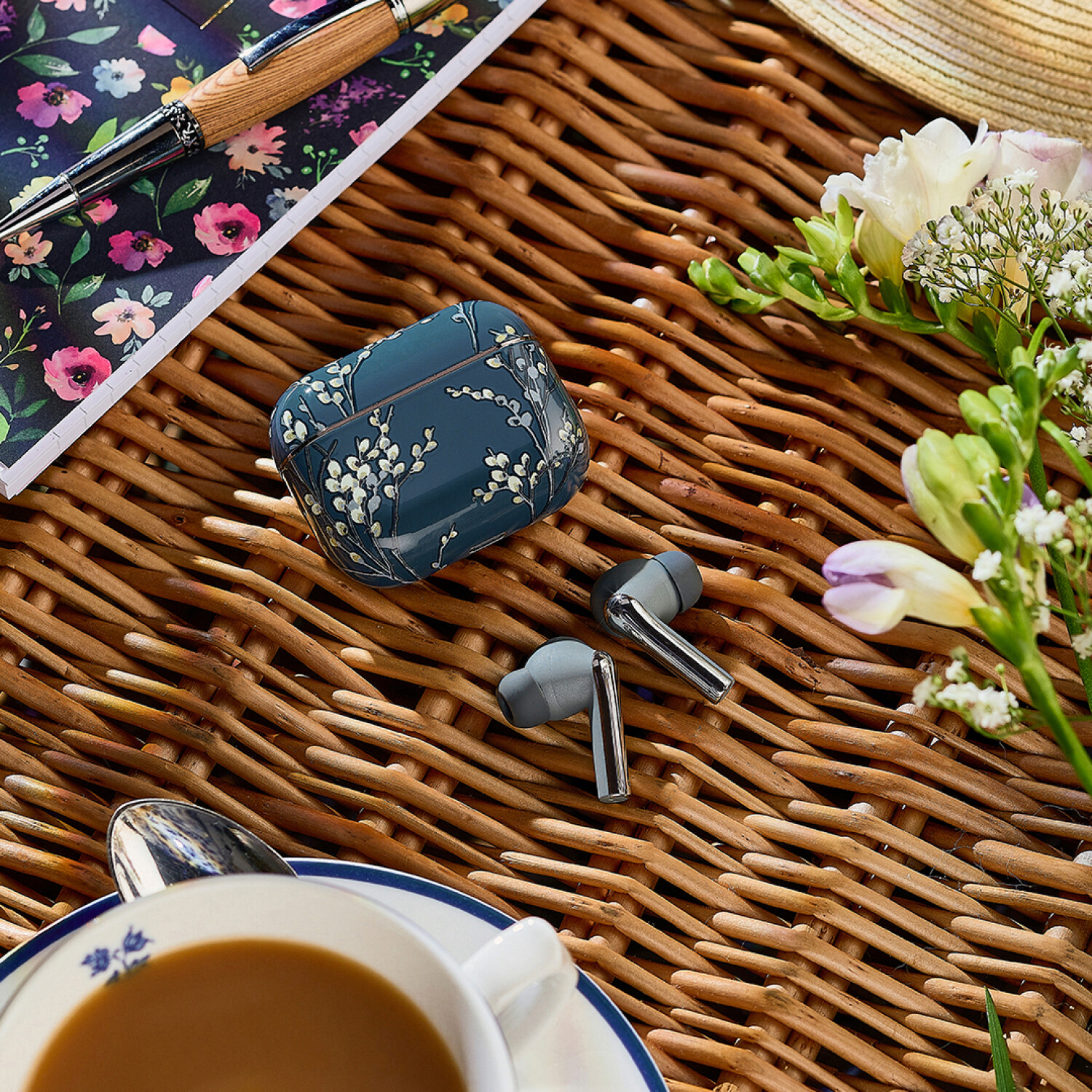 Ohrhörer, Hülle, Stift und Notizbuch auf einer Korboberfläche, mit Blumen und einer Kaffeetasse.