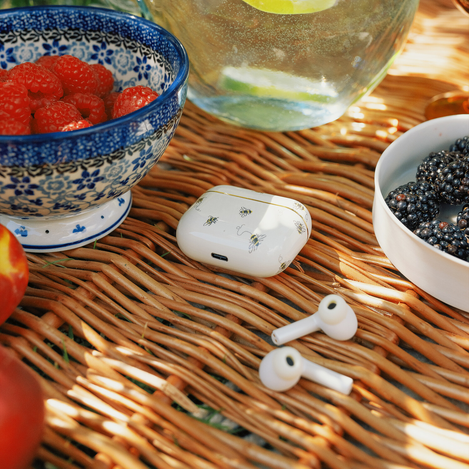 Eine Picknick-Szene mit Himbeeren, Brombeeren und einem weißen Etui mit Bienen auf einer gewebten Matte. Auch Ohrhörer sind vorhanden.
