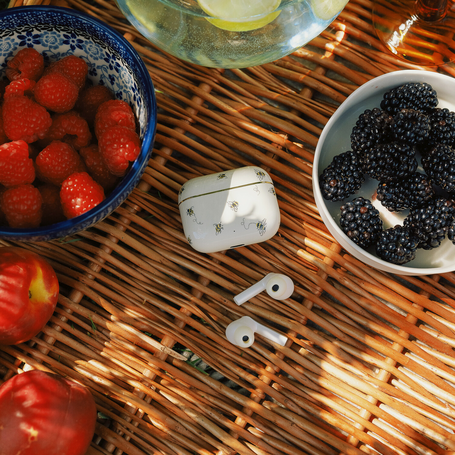 Eine Picknick-Szene mit Himbeeren, Brombeeren, Pfirsichen und Ohrhörern auf einer gewebten Matte. Es gibt ein Getränk und ein mit Bienen verziertes Etui.
