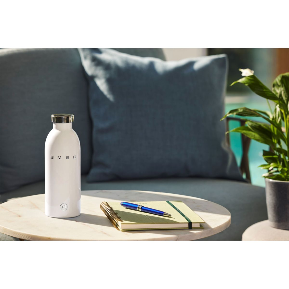 Eine weiße Smeg-Flasche, ein Notizbuch mit Stift und eine Pflanze stehen auf einem Tisch. Im Hintergrund sind eine Couch und Kissen zu sehen.