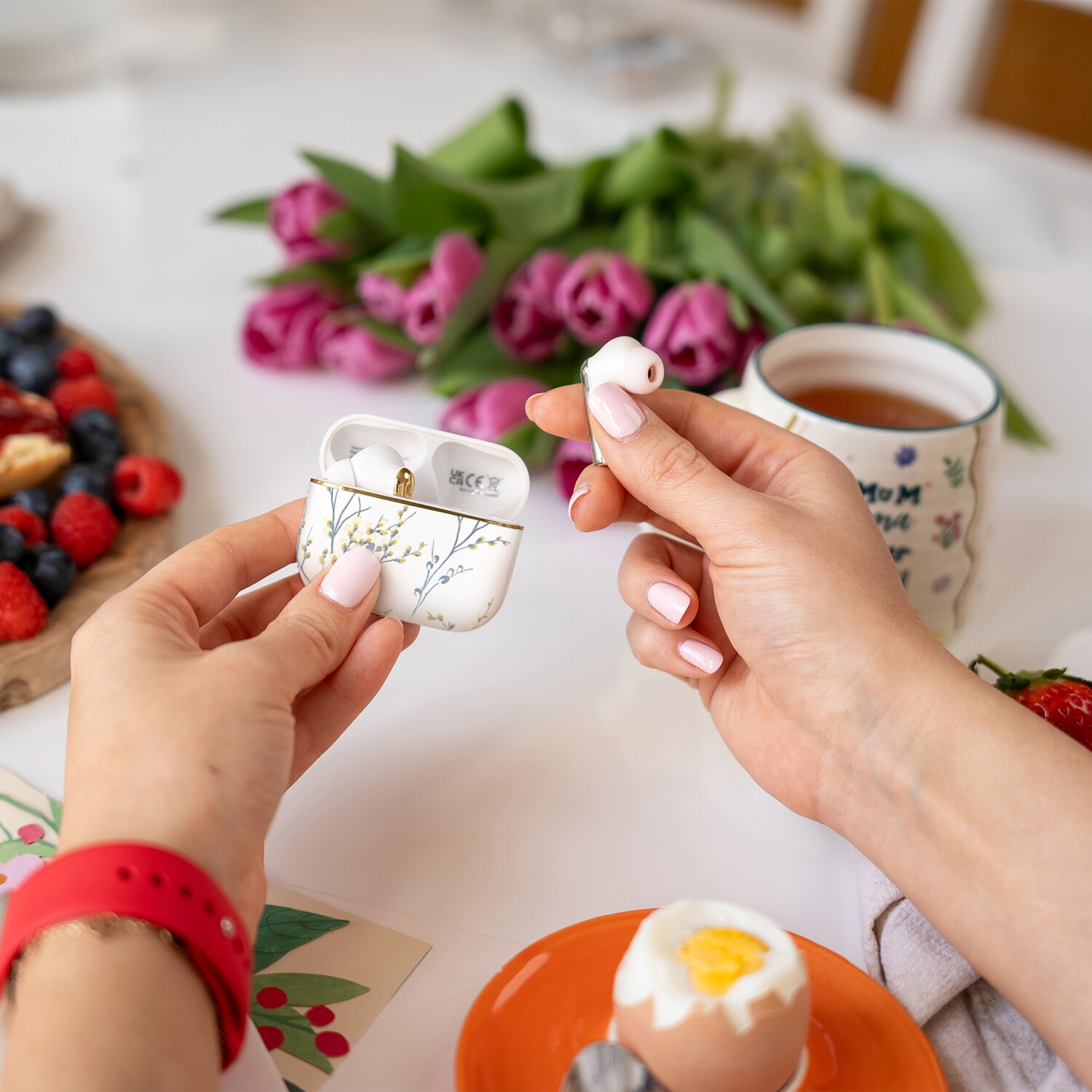 Hände halten Ohrhörer und Hülle mit Blumendesign, Frühstücksszene mit Früchten, Tulpen und einer Teetasse.