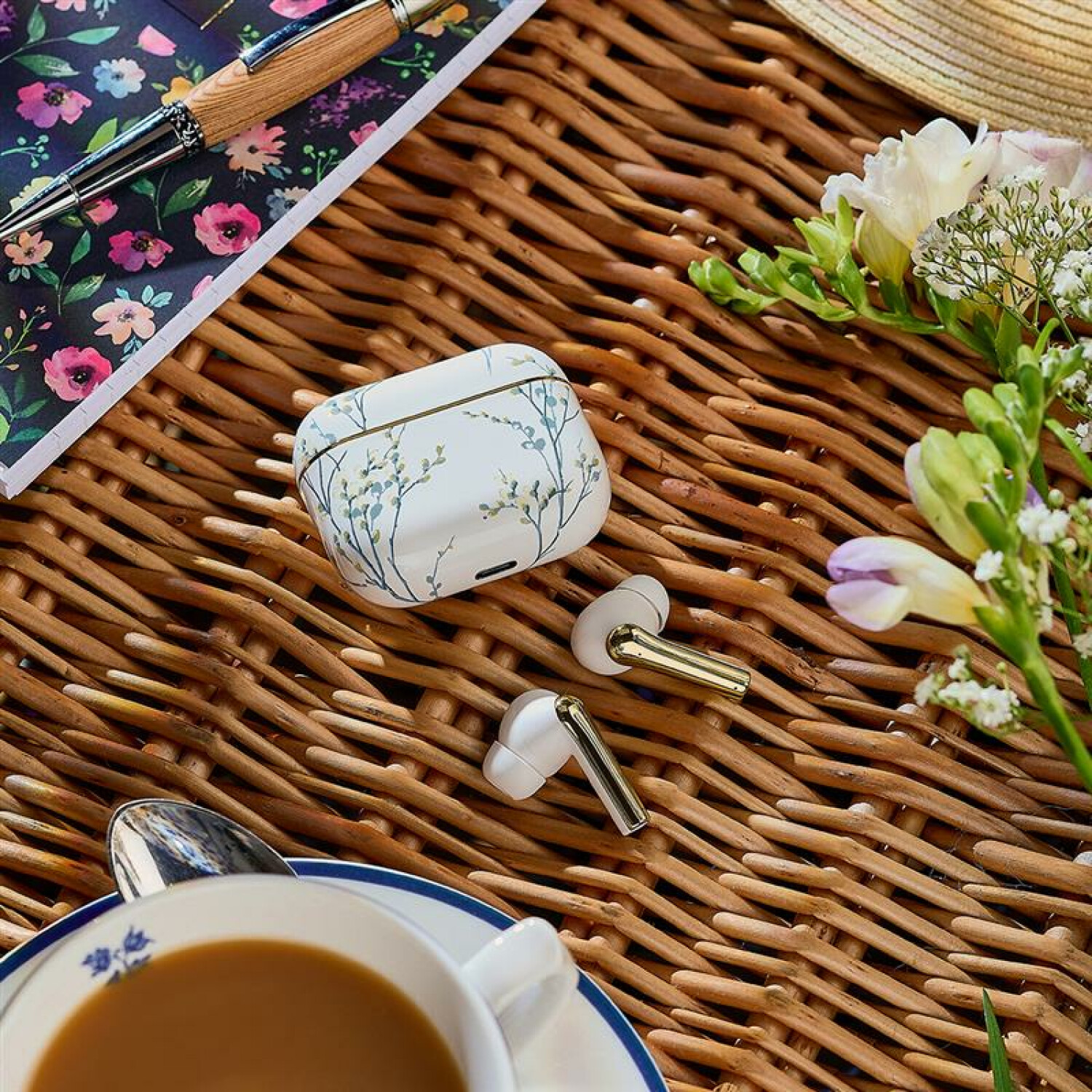 Weiße Ohrhörer, Etui, Blumen, Stift und Kaffeetasse auf einer Korboberfläche. Blumiges Notizbuch und goldfarbene Akzente.