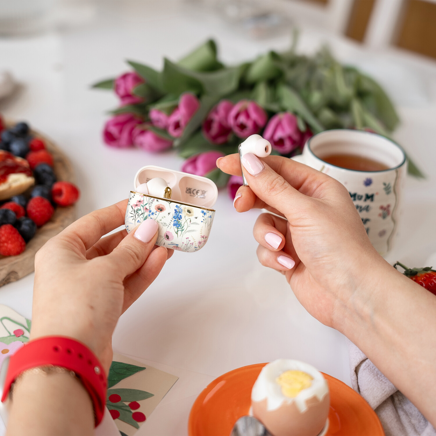 Hände halten Ohrhörer und Etui mit floralem Design, Frühstücksszene mit Beeren, Tulpen und einer Teetasse.