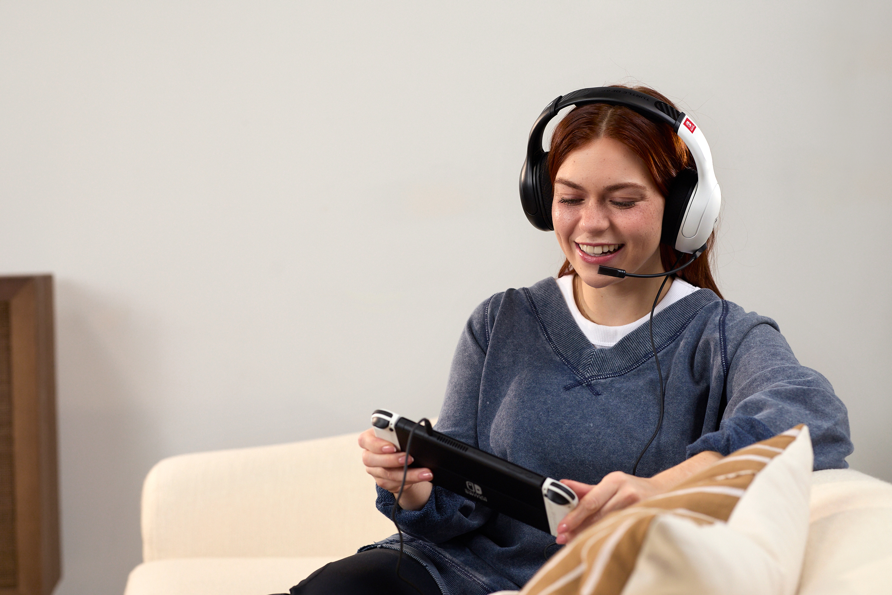 Ruda kobieta w słuchawkach gra w handhelda uśmiechając się na kanapie.