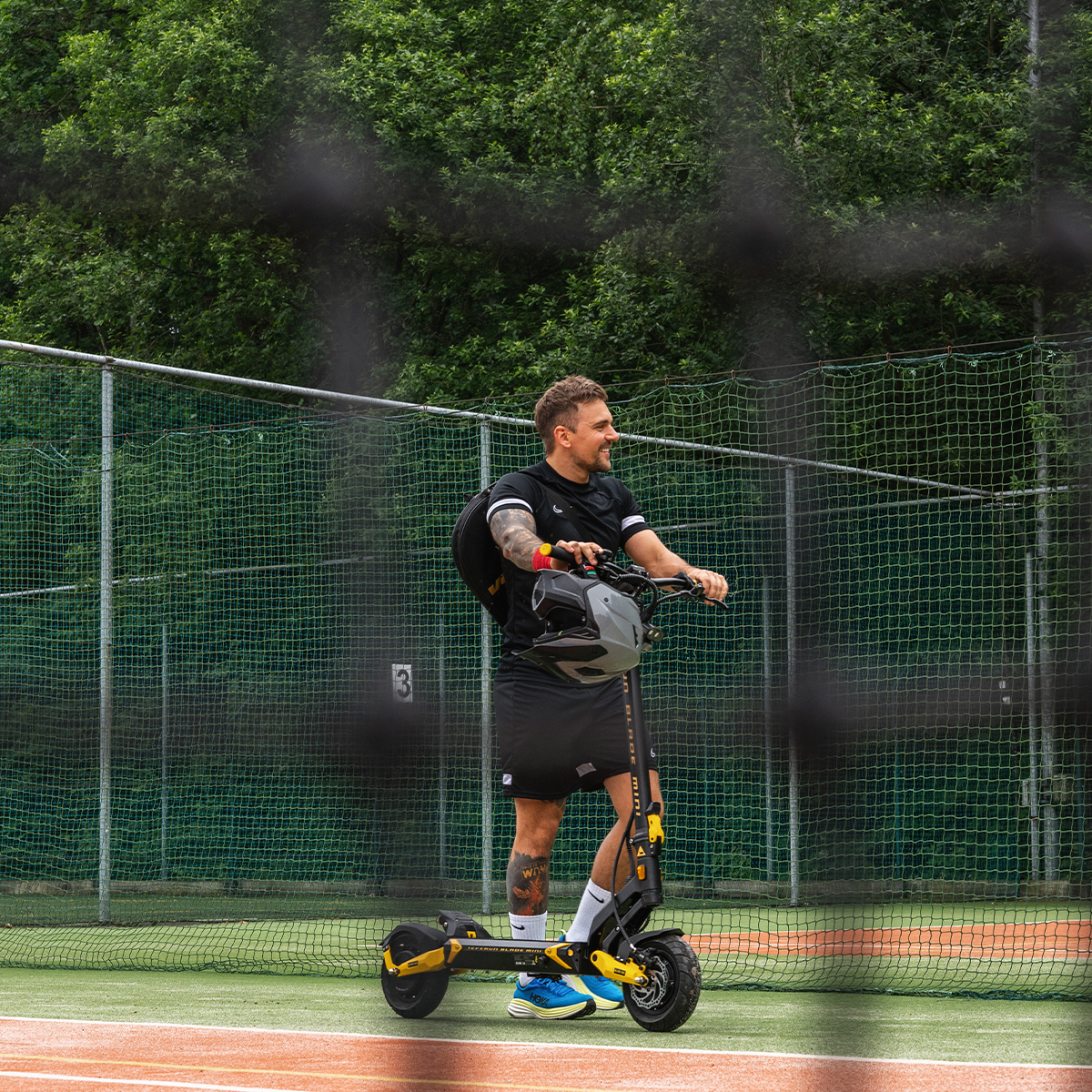 Mężczyzna stoi na korcie tenisowym, trzymając hulajnogę i kask. Ma tatuaże i się uśmiecha. Zielona siatka i drzewa w tle.