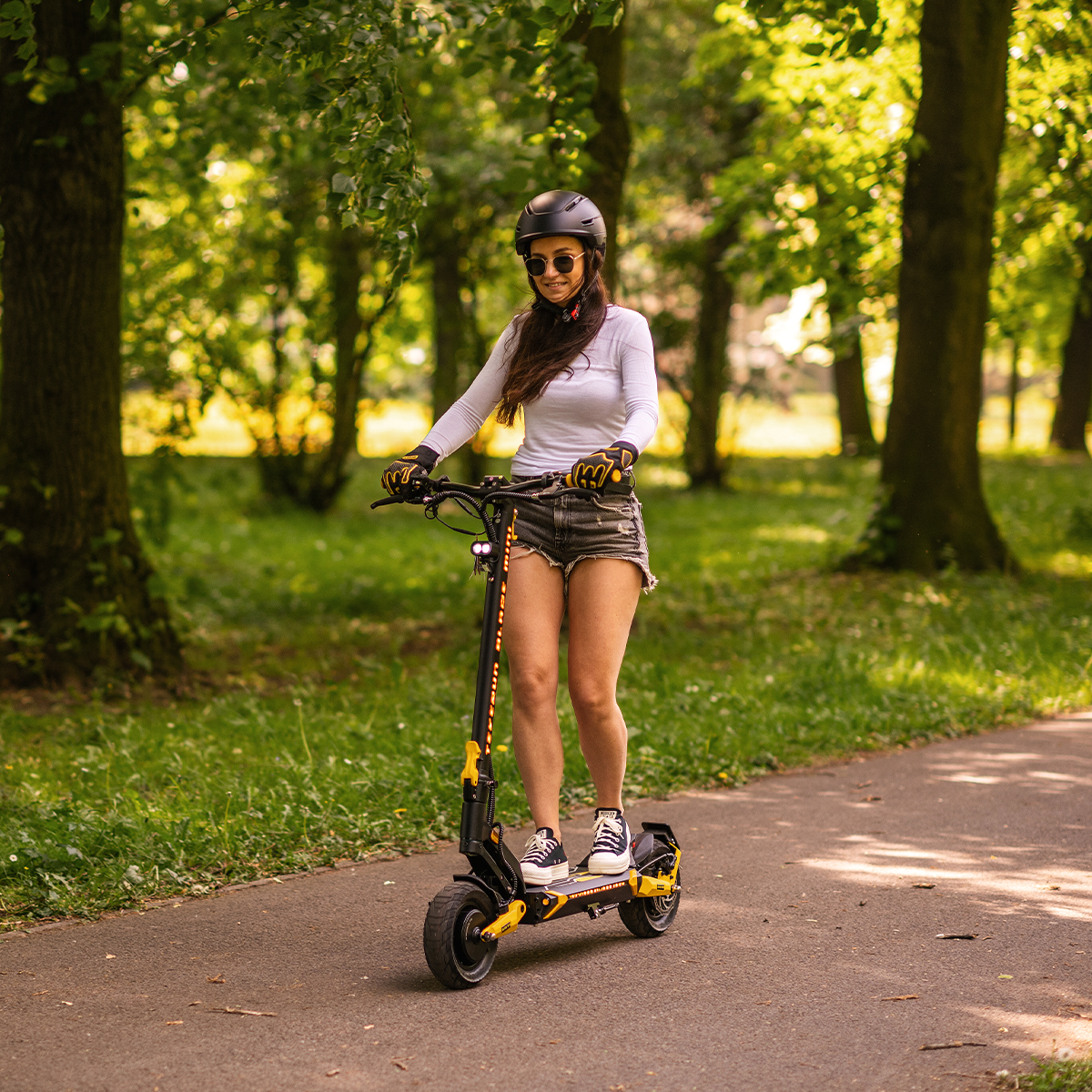 Kobieta jedzie na hulajnodze elektrycznej ścieżką w parku. Ma na sobie kask i szorty. W tle drzewa.