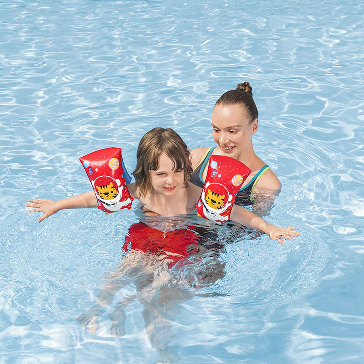 Eine Frau und ein Kind sind in einem Pool. Das Kind trägt rote Armbänder mit Tigerdesign und rote Shorts. Die Frau unterstützt das Kind.
