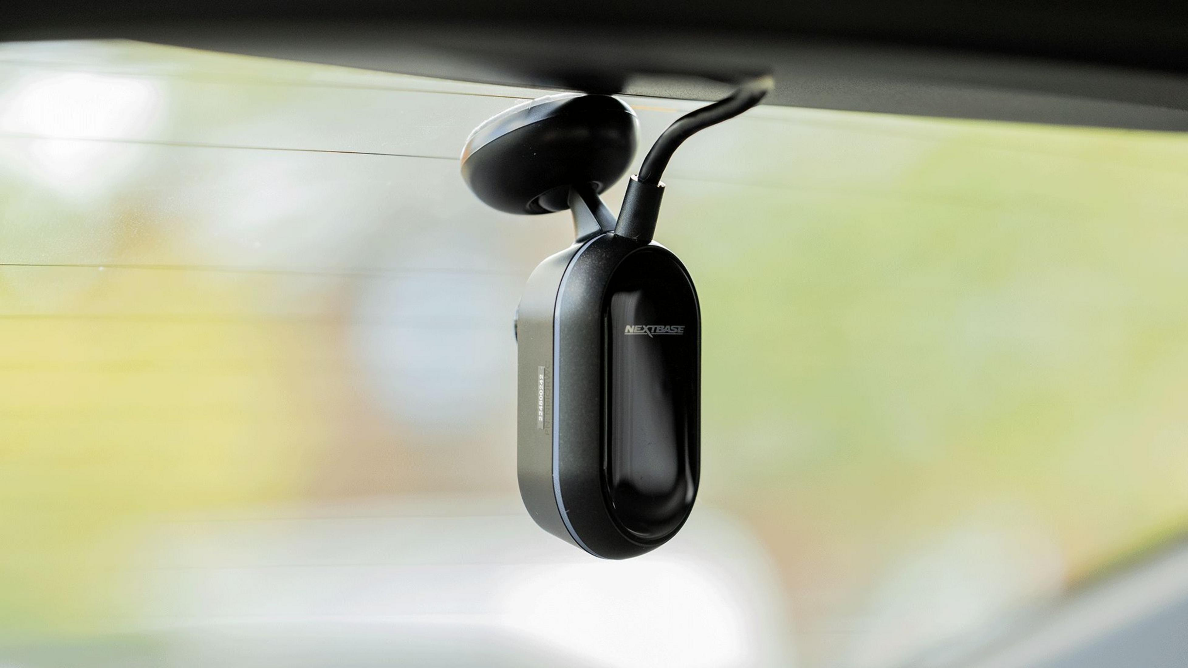Eine schwarze Dashcam an der Heckscheibe eines Autos befestigt. Das Nextbase-Logo ist sichtbar. Der Hintergrund ist grün und weiß verschwommen.