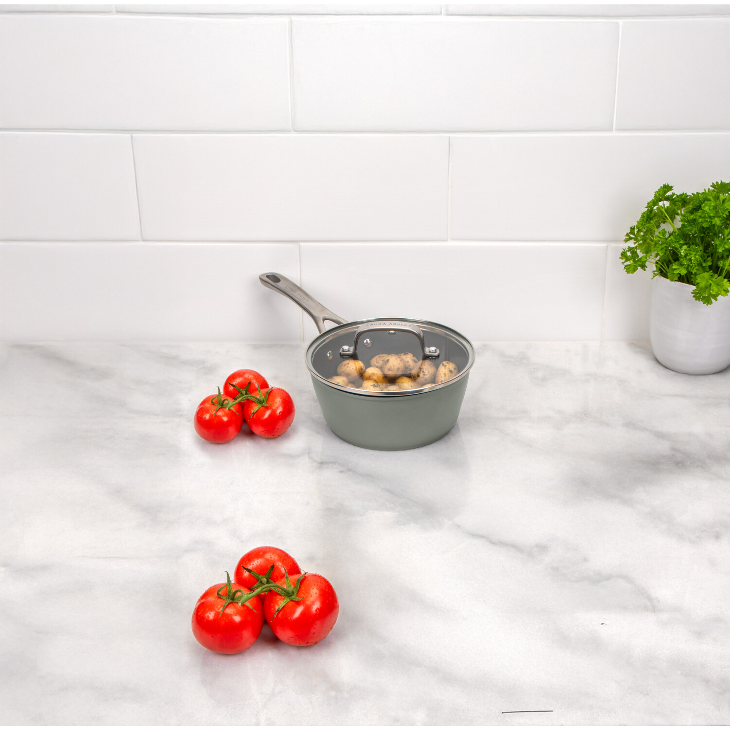 Eine Küchenszene mit einem Topf mit Kartoffeln, roten Tomaten und einer Topfpflanze auf einer Marmorarbeitsplatte. Weißer Fliesenhintergrund.