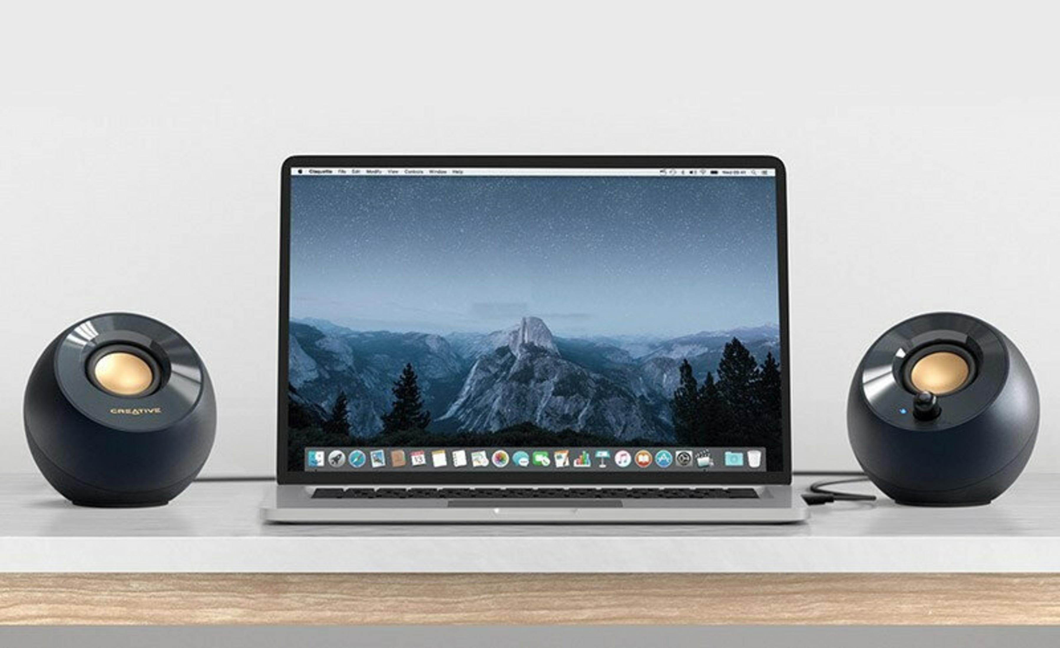Laptop z górskim krajobrazem na ekranie, otoczony dwoma czarnymi głośnikami ze złotymi akcentami, na białym biurku.