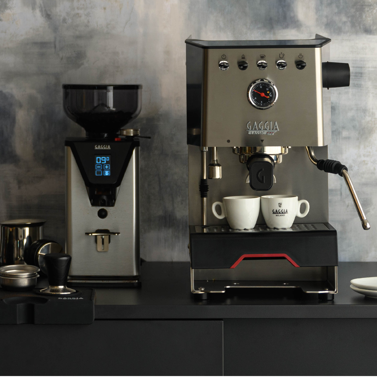 Kaffeemaschinen-Setup mit Mühle und Espressomaschine. Zwei weiße Tassen stehen bereit. Silberne und schwarze Geräte auf schwarzer Oberfläche.