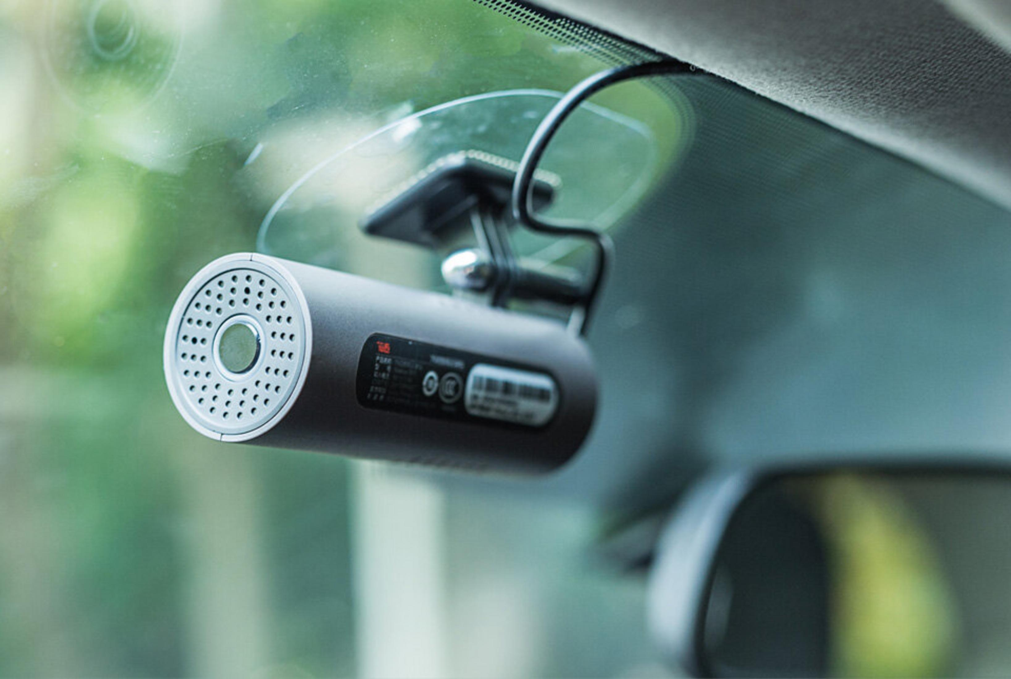 Eine Dashcam ist an der Windschutzscheibe eines Autos montiert. Sie ist zylindrisch und grau. Der Hintergrund zeigt das Innere des Autos.