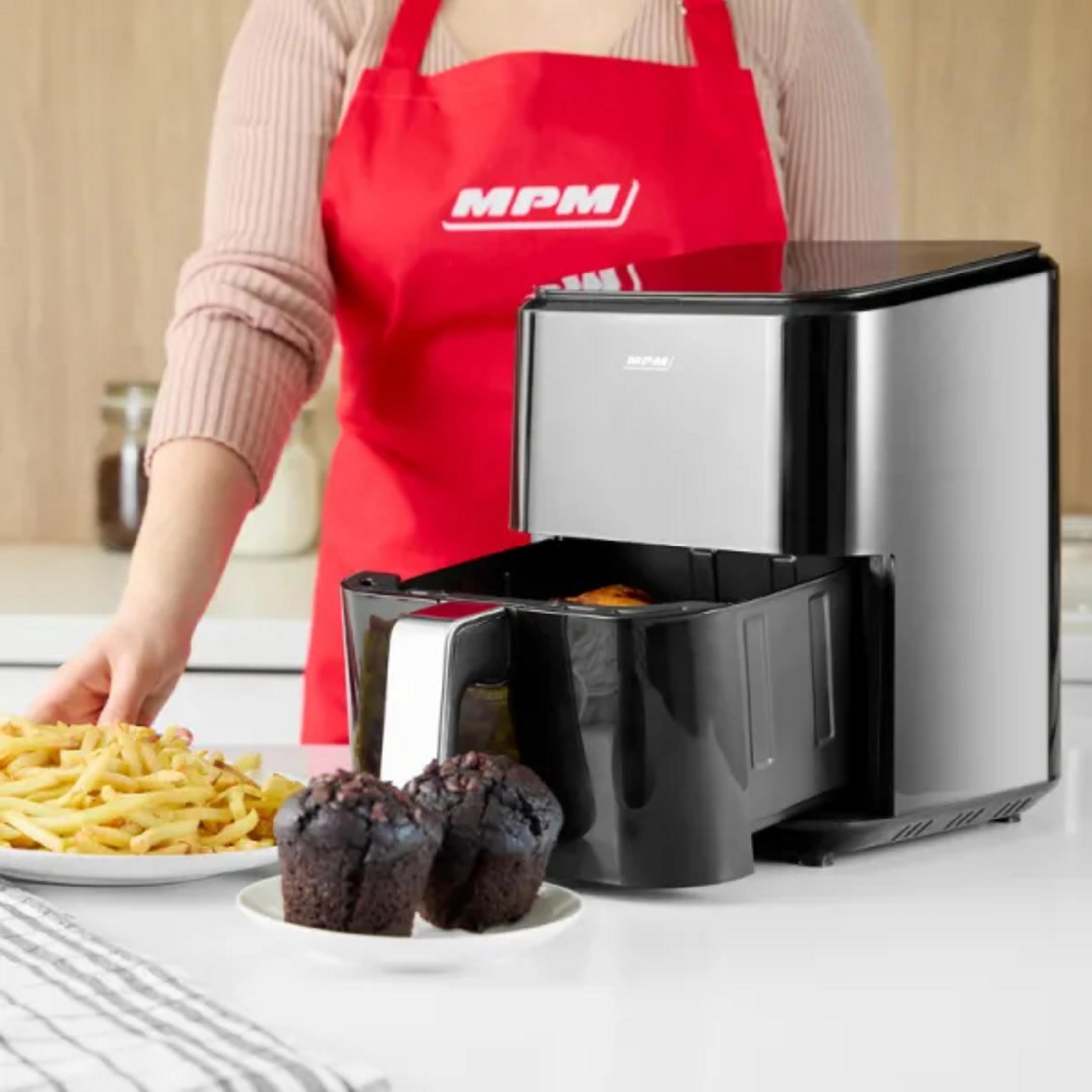 Una persona con un grembiule rosso è in piedi vicino a una friggitrice ad aria con muffin e patatine fritte su un tavolo.