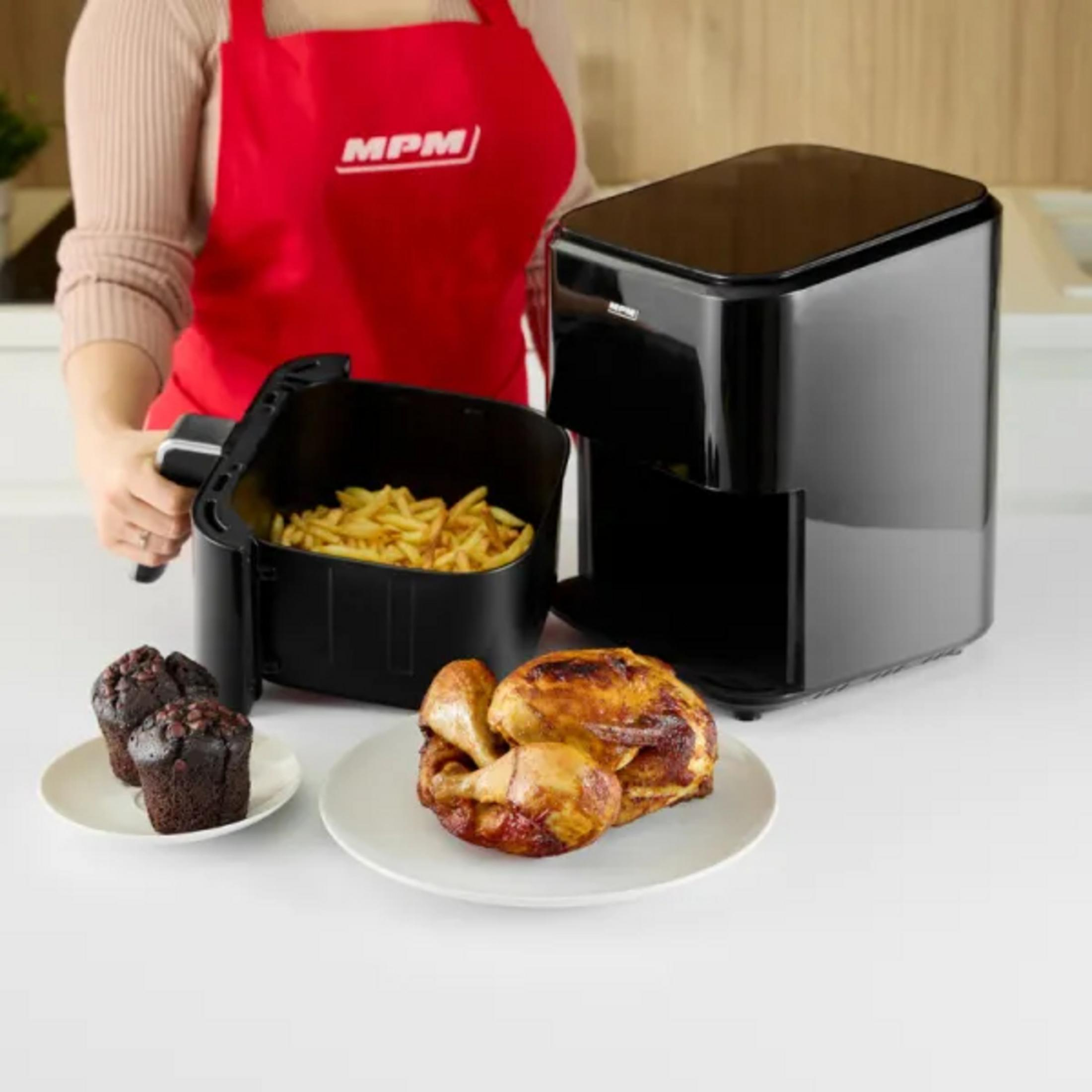 Una friggitrice ad aria con cibo, tra cui patatine fritte, muffin e pollo. Una persona con un grembiule rosso tiene il cestello della friggitrice.