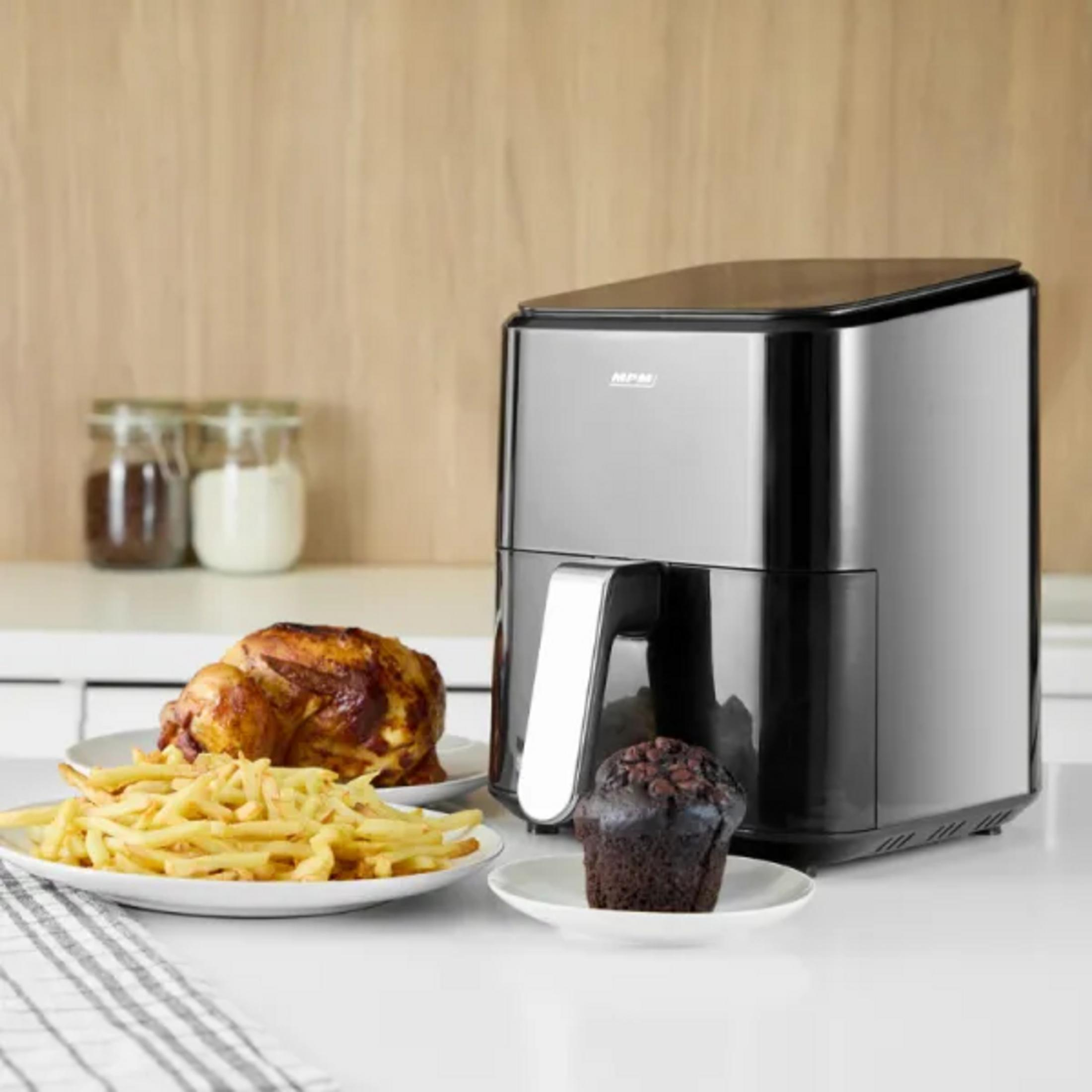 Una friggitrice ad aria con cibo come pollo, patatine fritte e un muffin su una superficie bianca. Lo sfondo ha barattoli e un muro di legno.