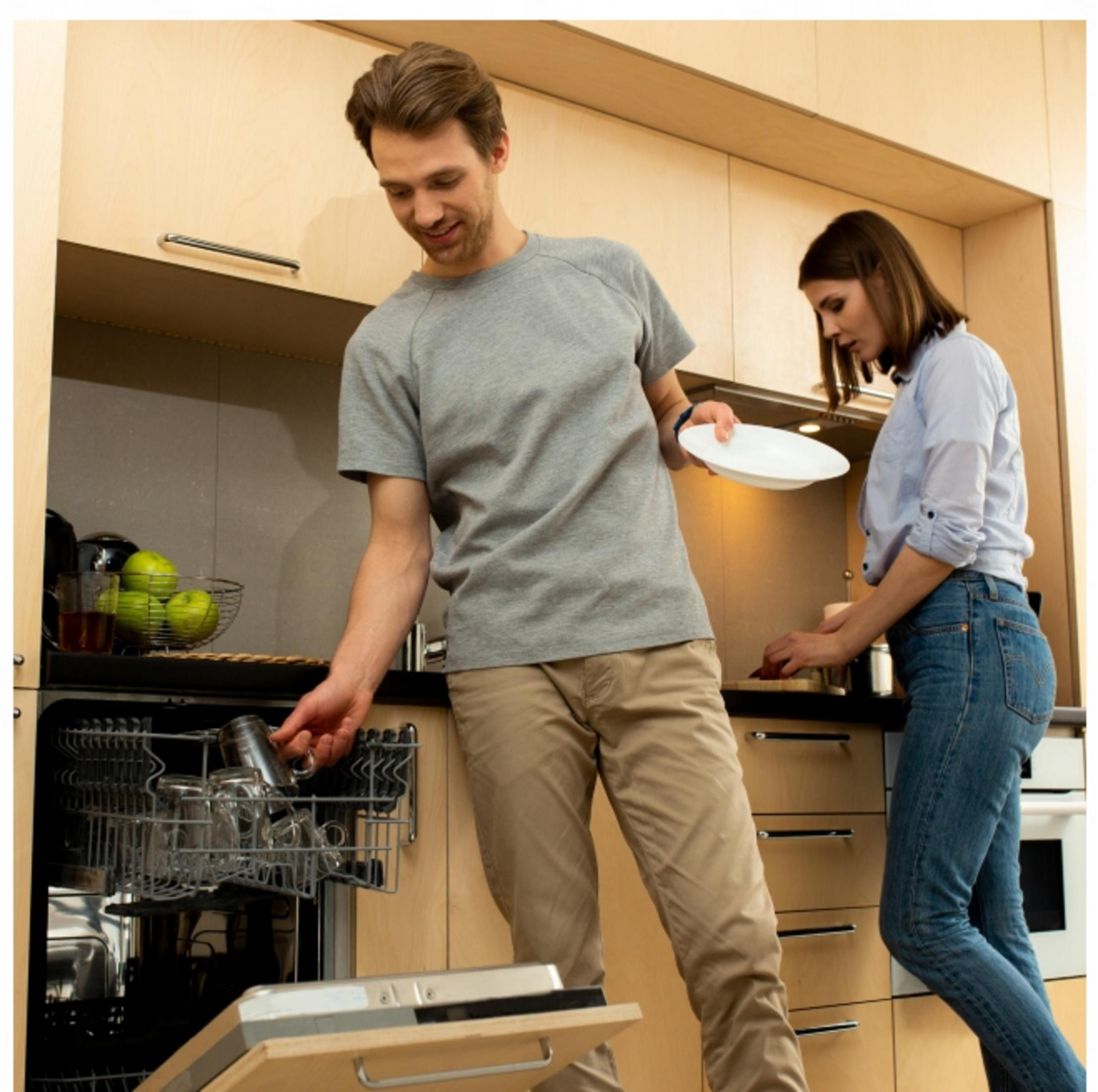 Mężczyzna wkłada szklanki do zmywarki, a kobieta trzyma talerz. W tle szafki kuchenne.