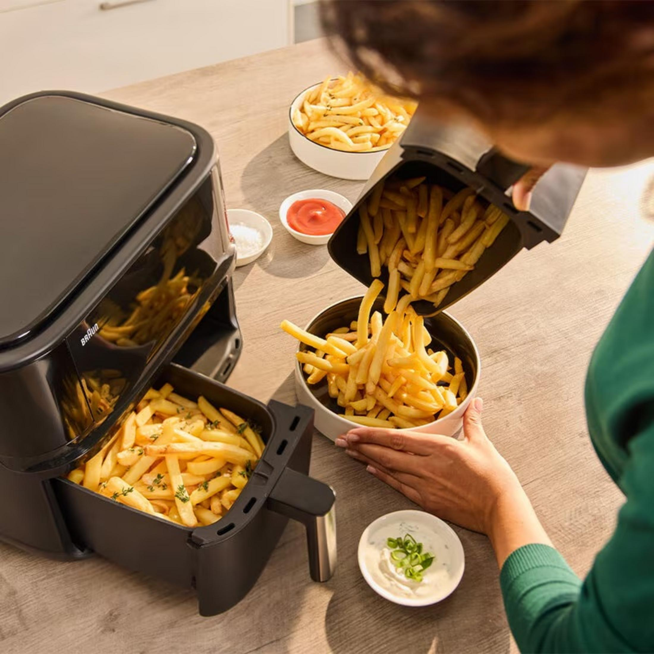 Een persoon giet frietjes uit een airfryer in een kom. Ketchup, zout en saus staan op tafel.